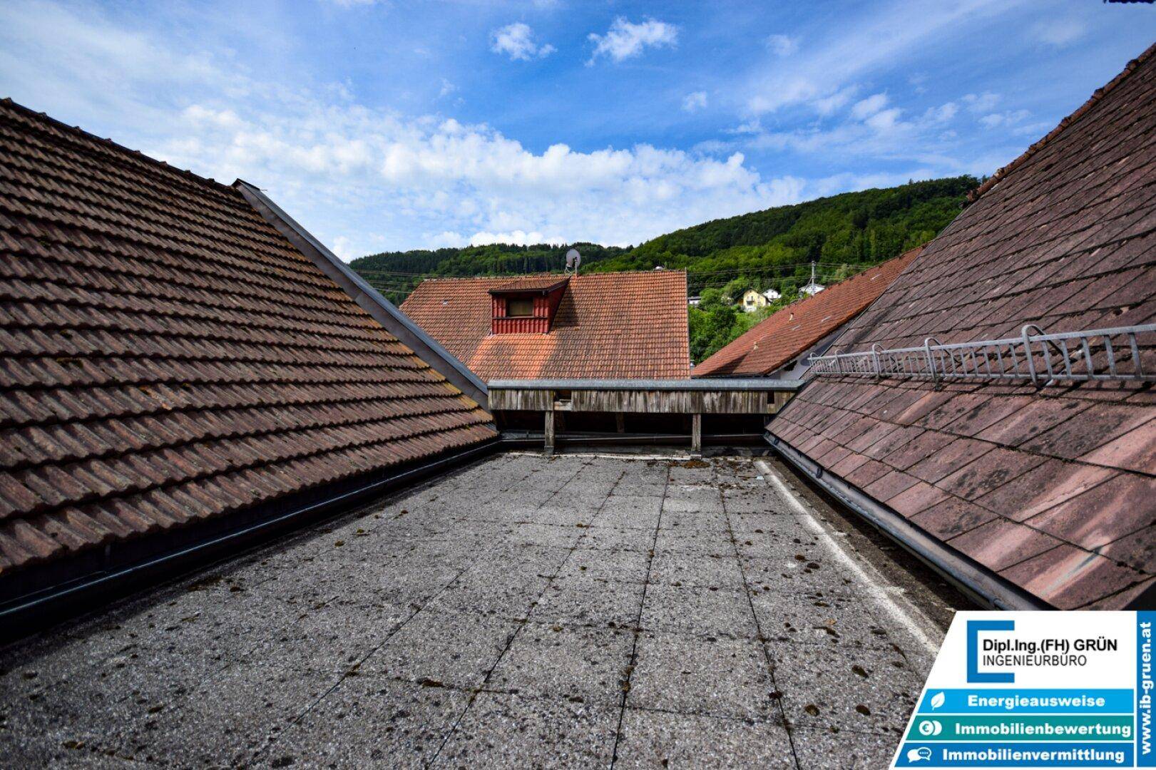 Blick über die Dächer alter Gebäude mit Ziegeln und einem Flachdach, umgeben von grünen Hügeln.