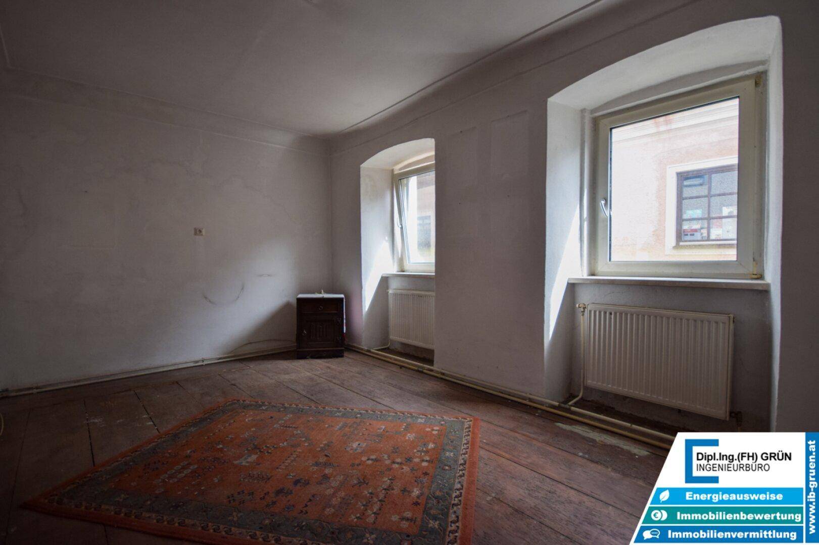 Heller Raum mit breiten Holzdielen, Heizkörpern unter den Fenstern und einem Teppich.