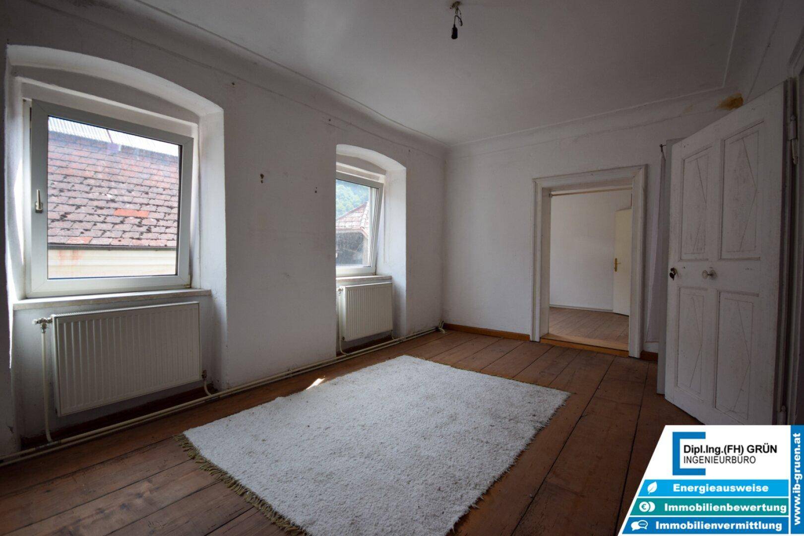 Schlafzimmer mit Holzdielenboden, zwei Fenstern und einem weißen Teppich.