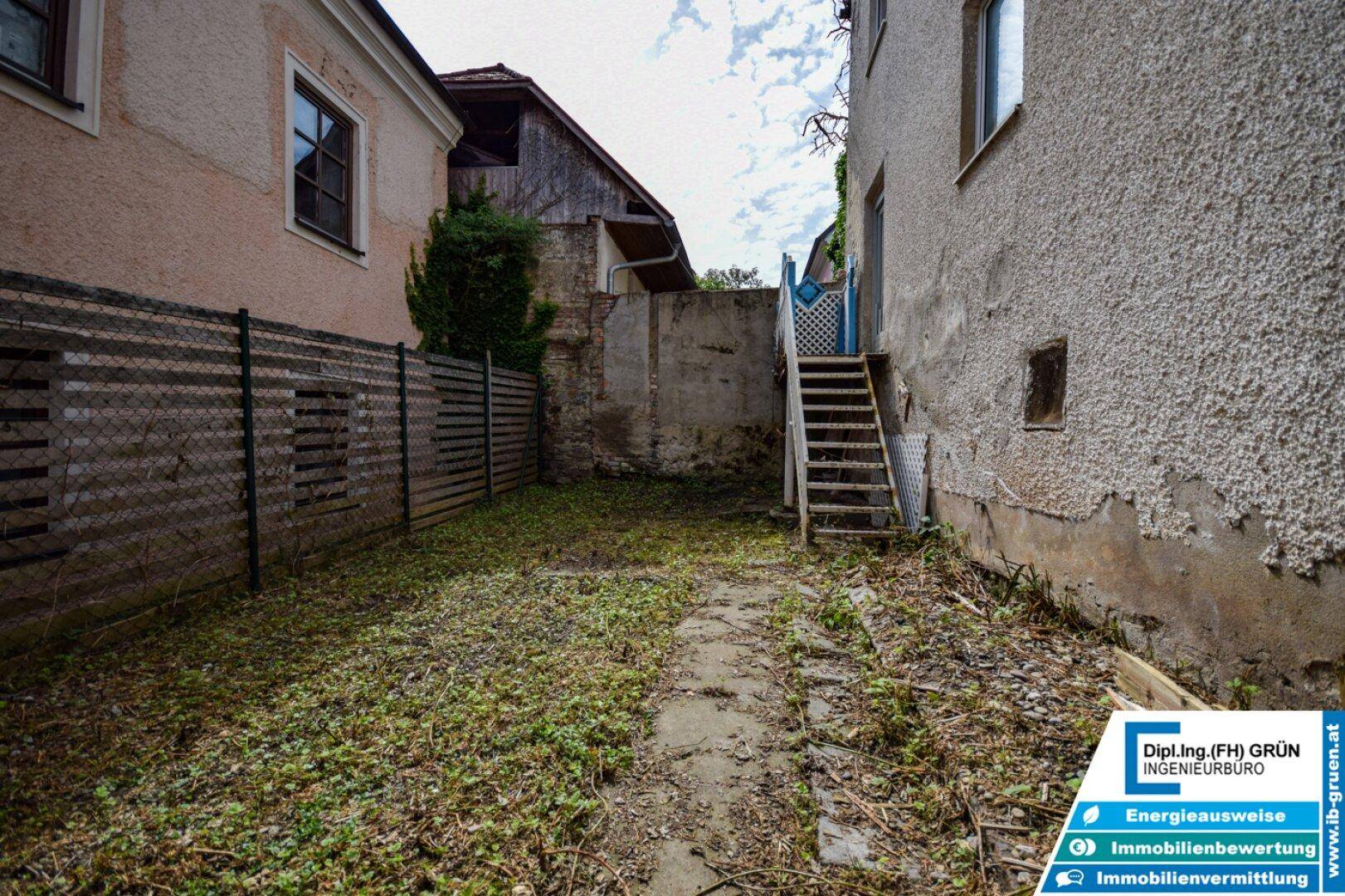Ungepflegter Hinterhof mit Gartenfläche, alten Gebäuden und einer Holztreppe.