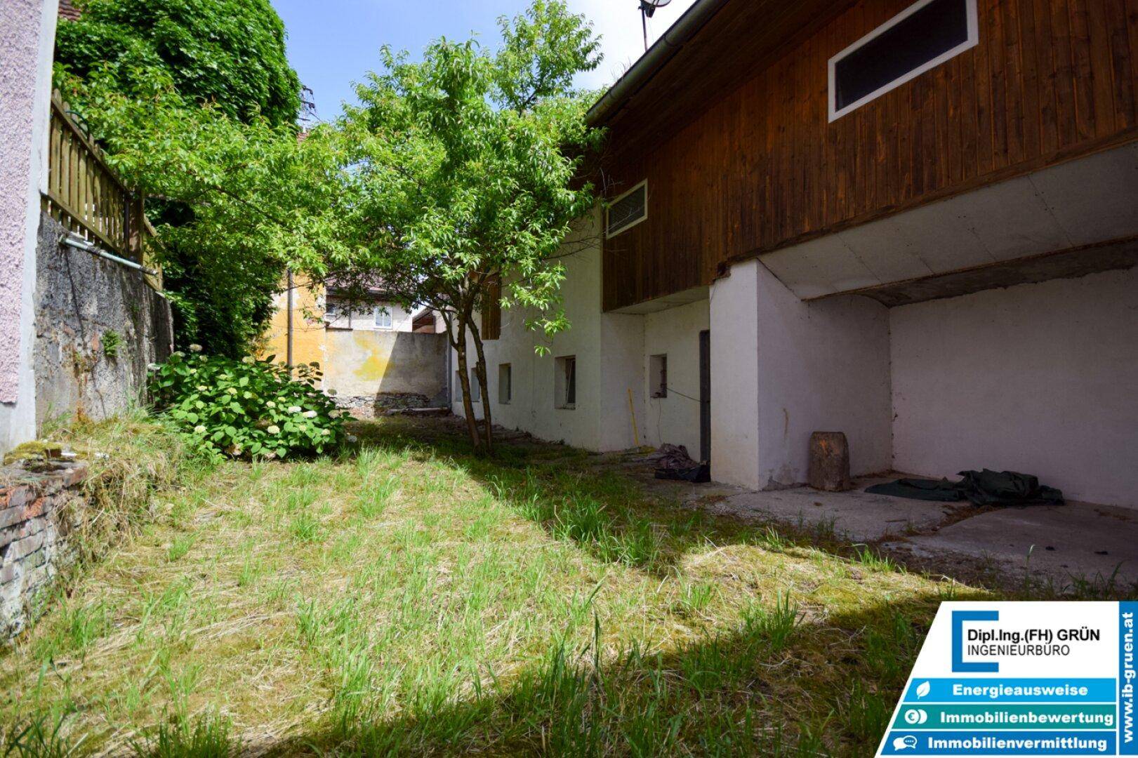 Ungepflegter Gartenbereich mit Gras, Bäumen und der Rückseite eines Gebäudes mit Holzelementen.