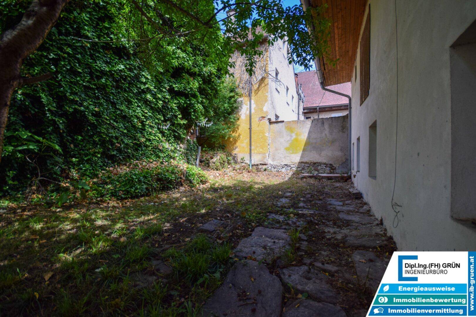 Verwilderter Gartenbereich mit Steinplatten, Efeu an der Wand und einem alten Gebäude im Hintergrund.