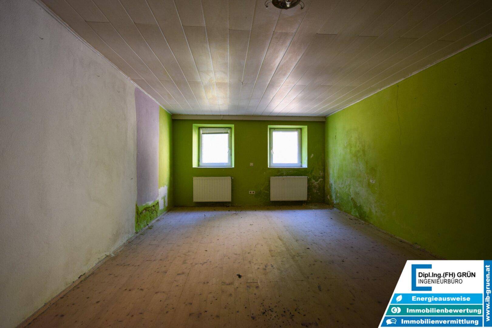 Leeres Zimmer mit grünen Wänden, Holzdecke und zwei Fenstern, die viel Licht spenden.