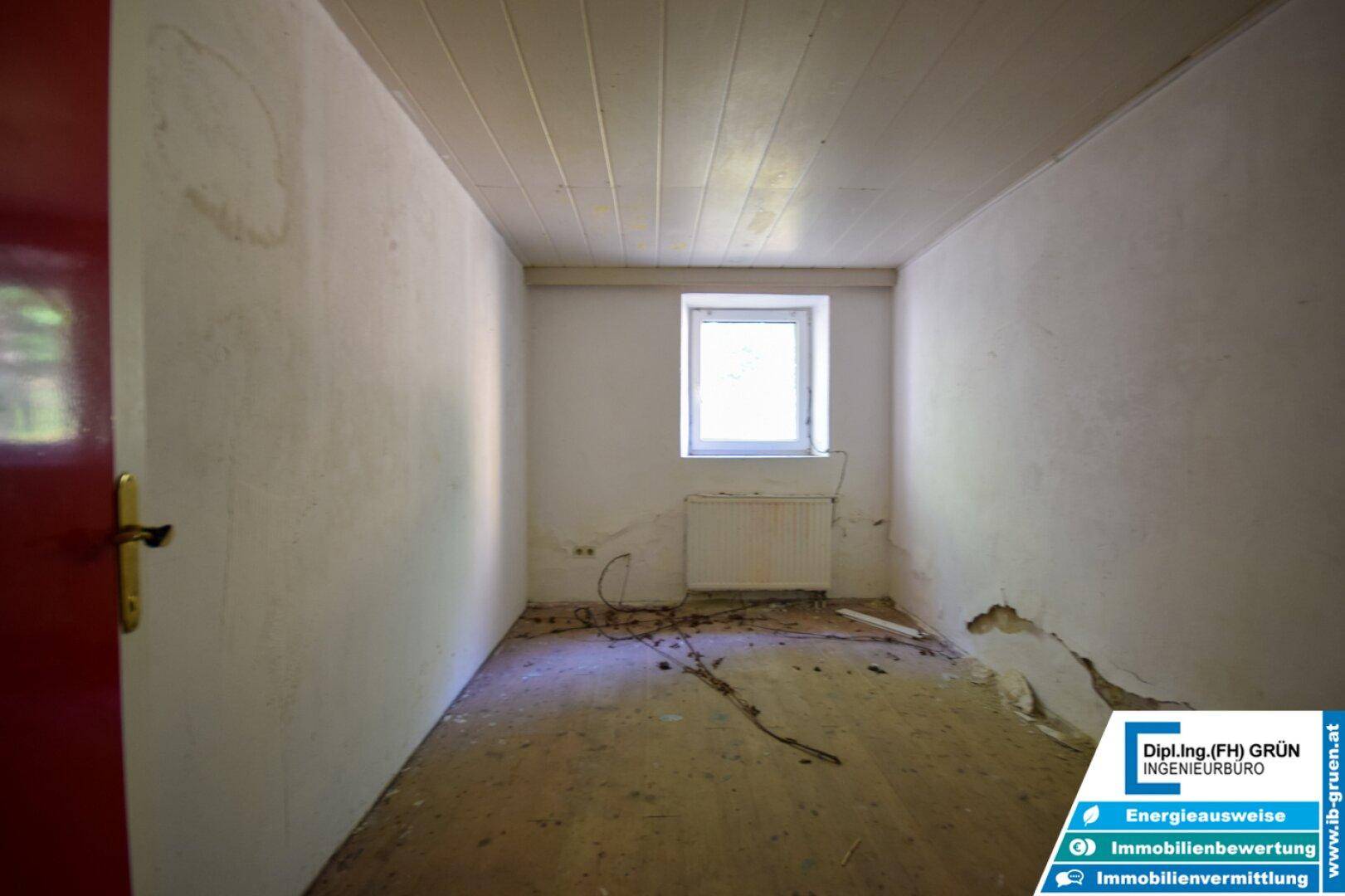 Kleiner, leerer Raum mit einem Fenster, Heizkörper und sichtbaren Abnutzungsspuren an den Wänden.