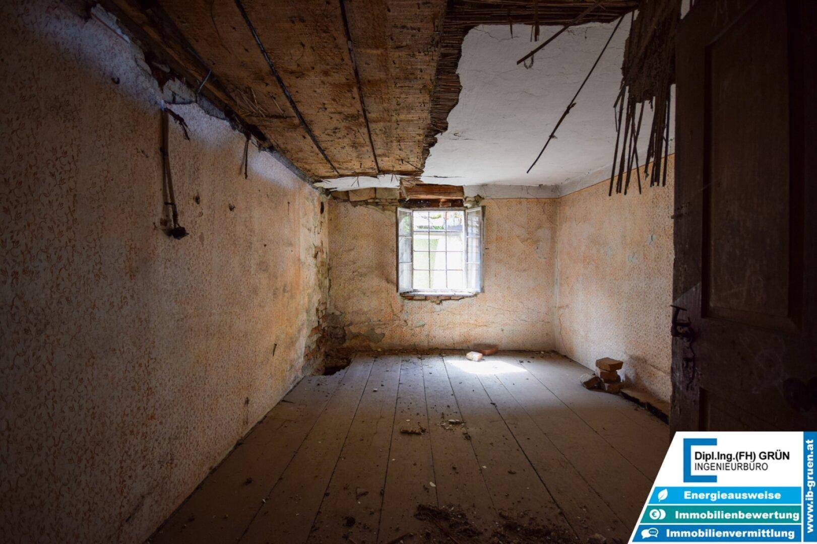 Renovierungsbedürftiger Raum mit freiliegenden Deckenbalken, einem Fenster und alten Holzdielen.