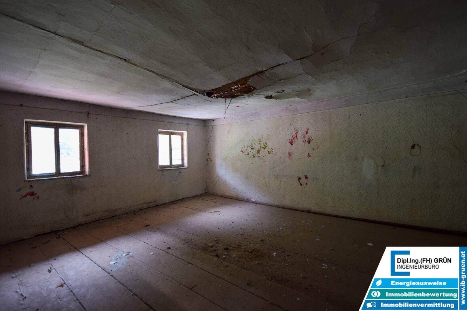 Alter Raum mit zwei Fenstern, Holzdielenboden und beschädigter Decke, die Renovierung erfordert.