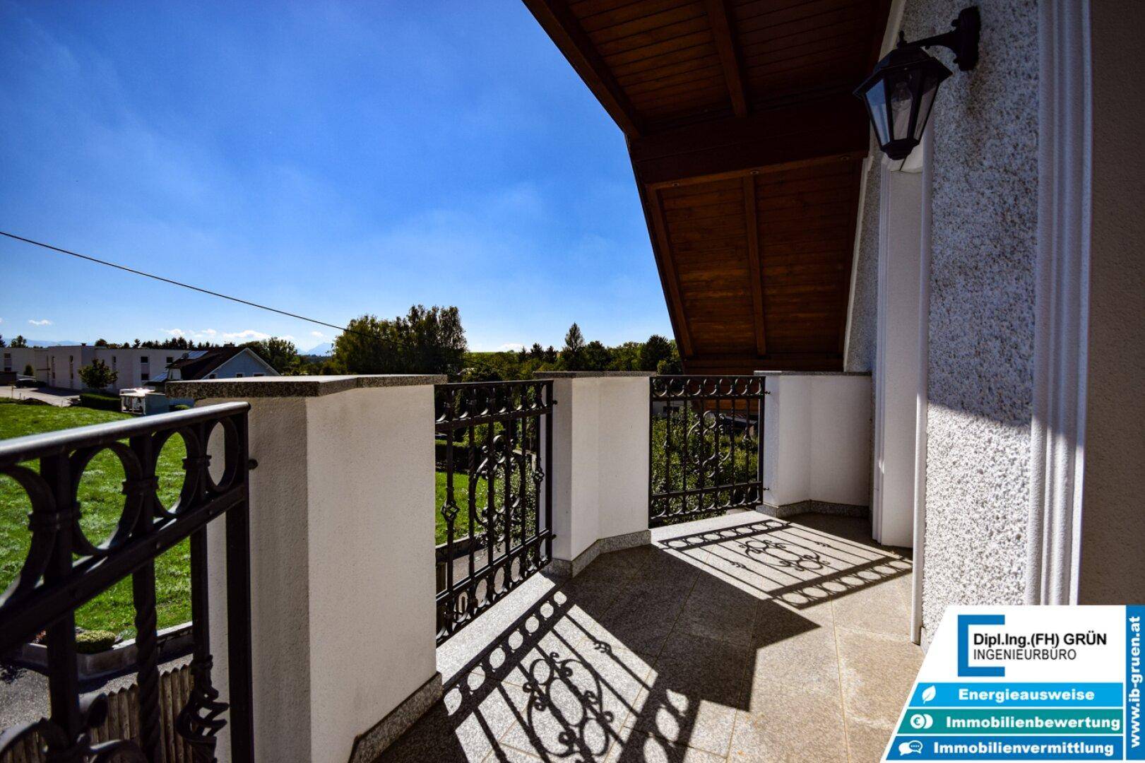 Balkon mit schmiedeeisernem Geländer und weitem Blick ins Grüne unter blauem Himmel.