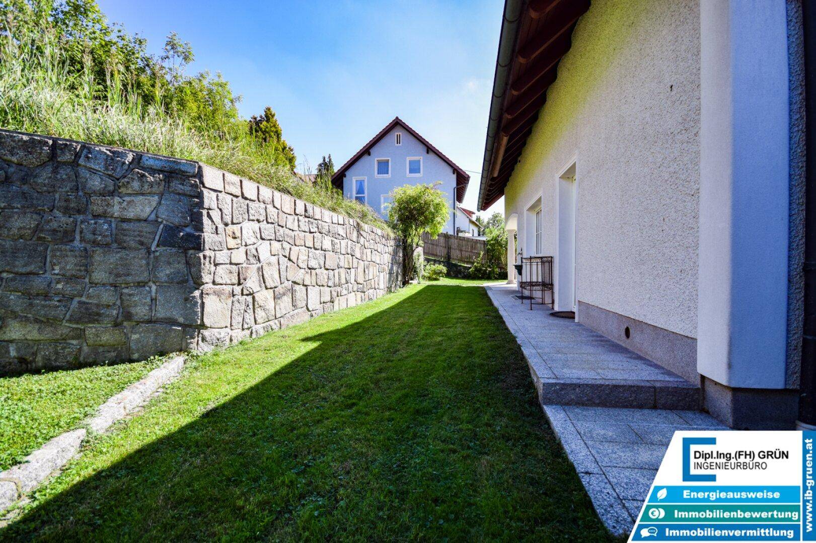 Langer Rasenstreifen entlang des Hauses mit Steinmauer und Blick auf Nachbarhaus.