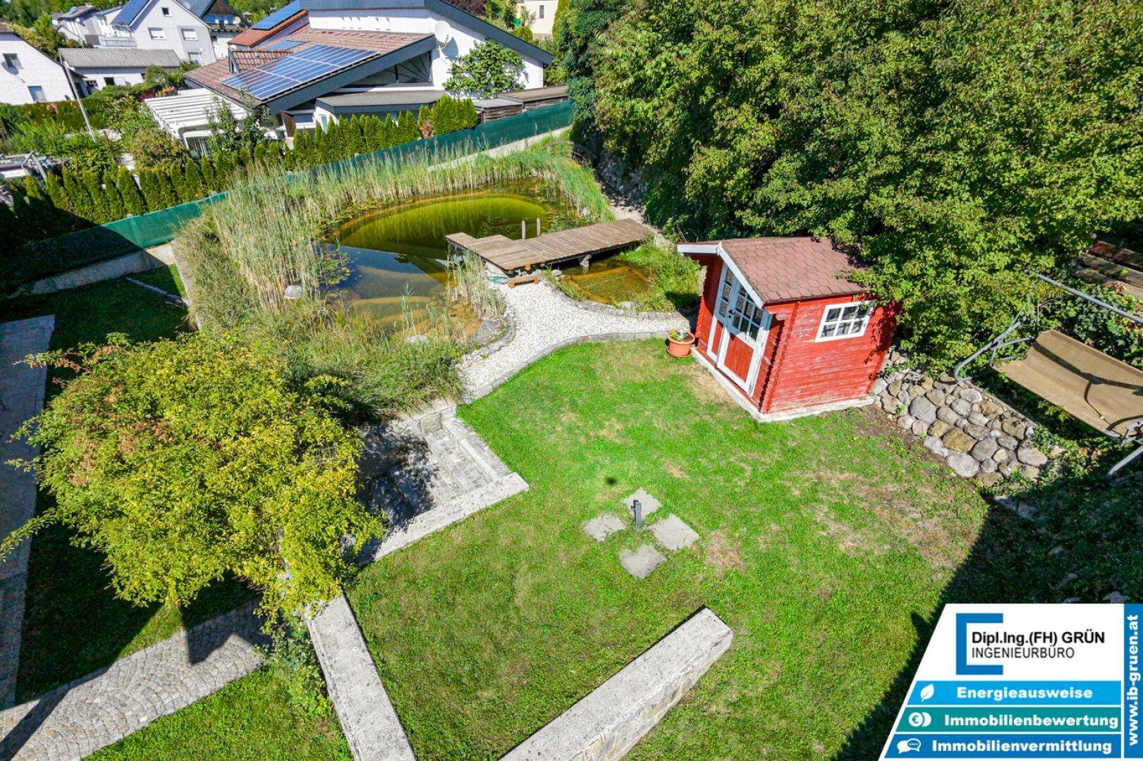 Gartenanlage von oben mit Teich, Holzsteg, rotem Gartenhaus und Rasenflächen.