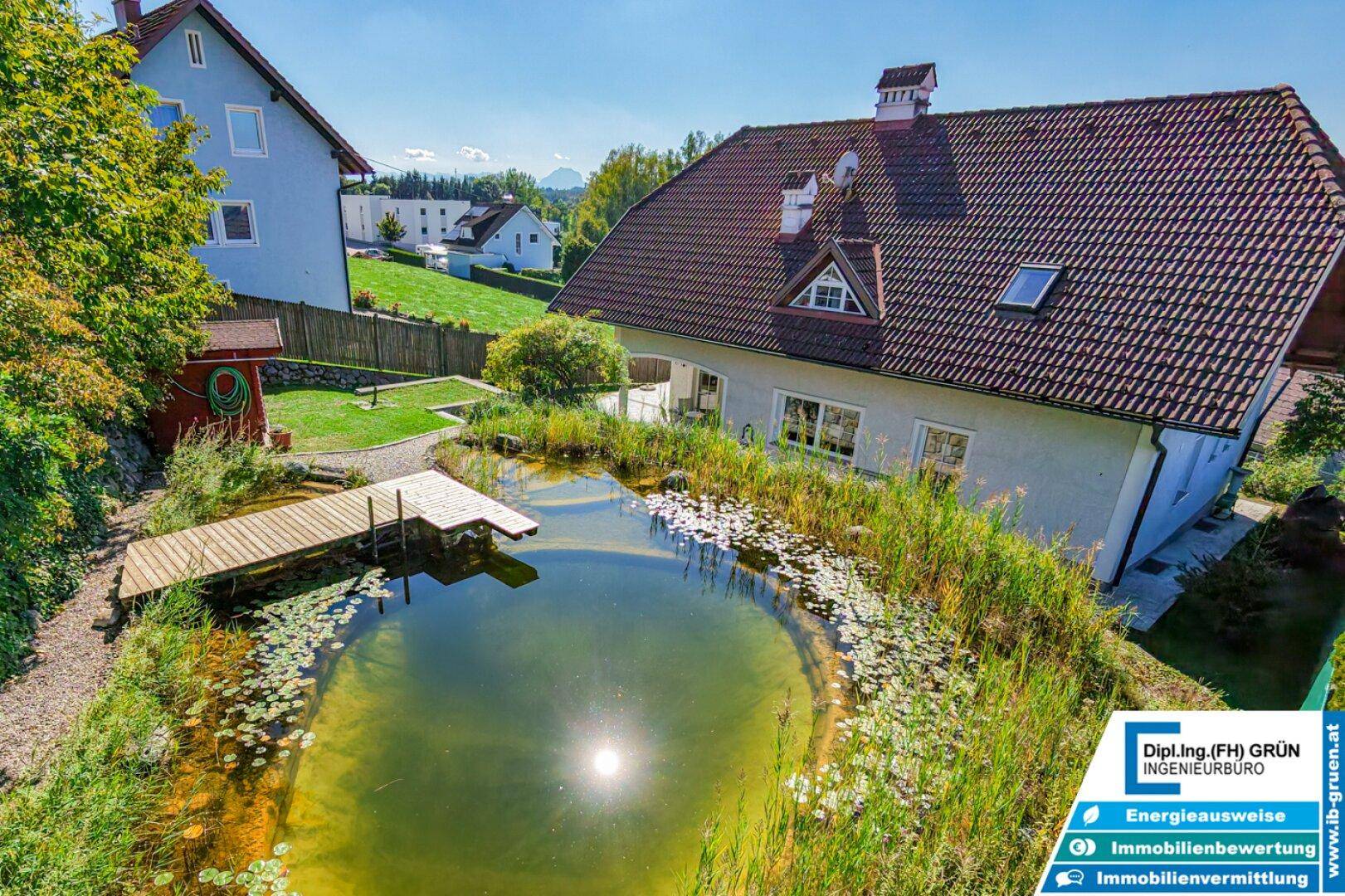 Hausansicht über einen Gartenteich mit Holzsteg und üppiger Uferbepflanzung.