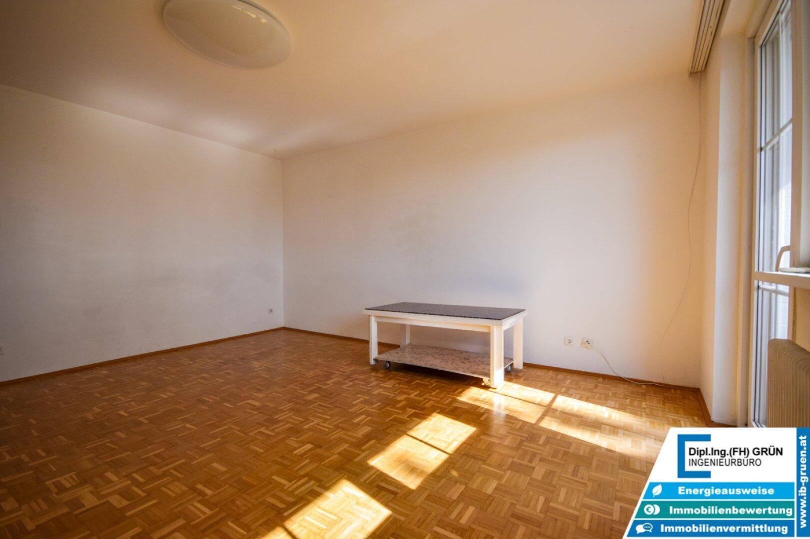 Leerer Raum mit Parkettboden, einem kleinen Tisch und viel Tageslicht.