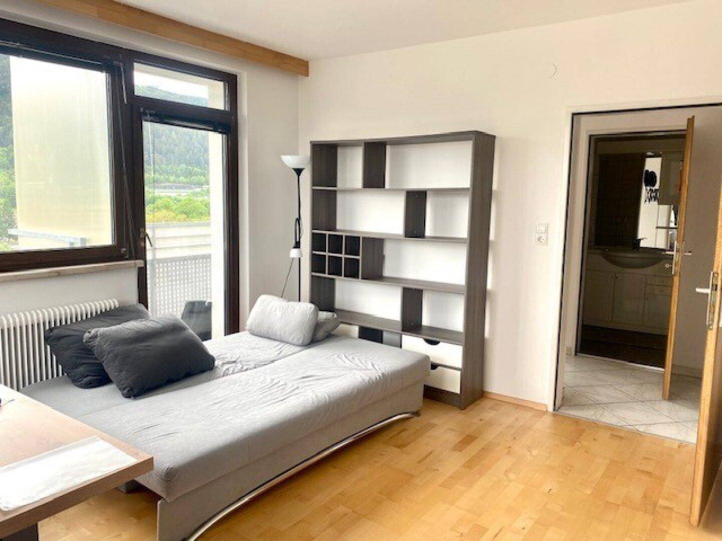 Helles Zimmer mit Schlafsofa, Bücherregal und Balkonzugang, mit Blick auf die Berge.