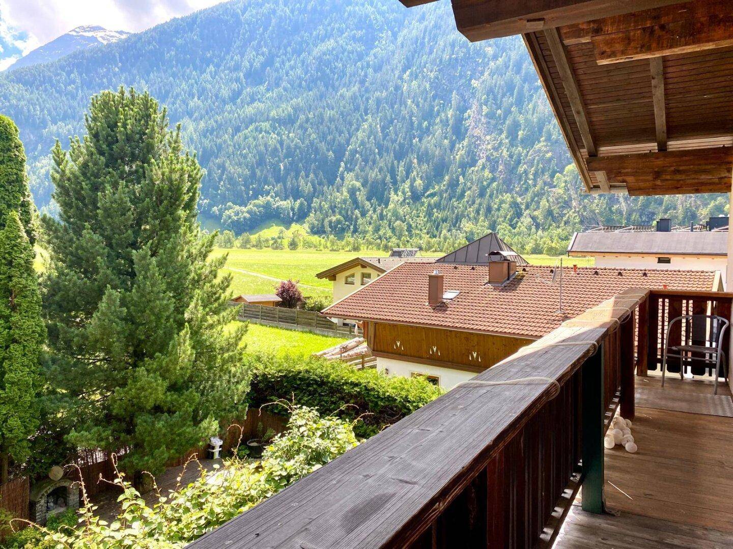 Panoramablick von einem Balkon auf die Alpenlandschaft mit bewaldeten Bergen und traditionellen Häusern.