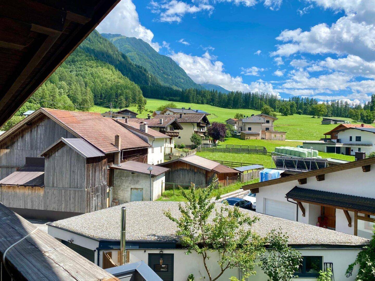 Weitblick von einem Balkon über ein idyllisches Dorf mit grünen Wiesen und majestätischen Bergen im Hintergrund.