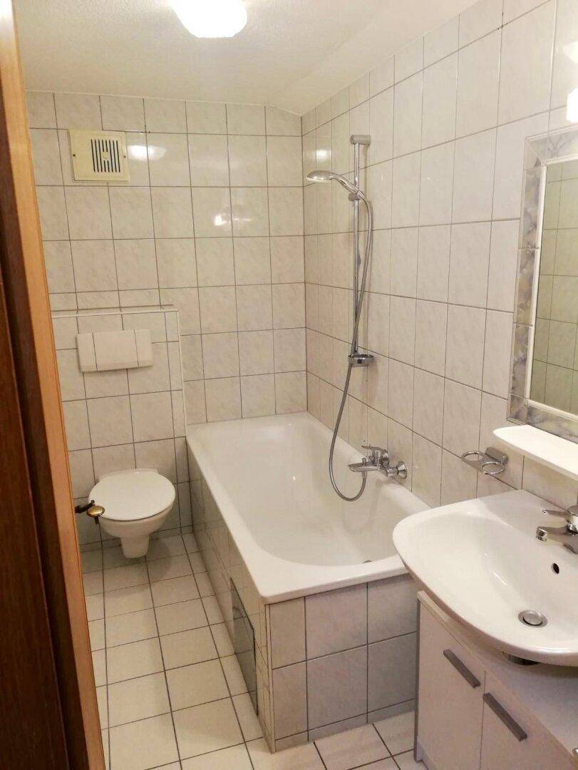 Funktionales Badezimmer mit weißen Fliesen, Badewanne mit Duschfunktion, WC und Waschbecken.