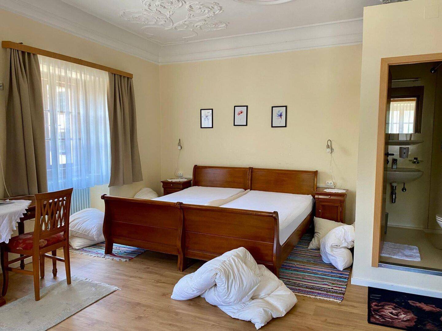 Helles Schlafzimmer mit traditionellem Holzbett, Stuckdecke und Blick auf die Umgebung durch große Fenster.