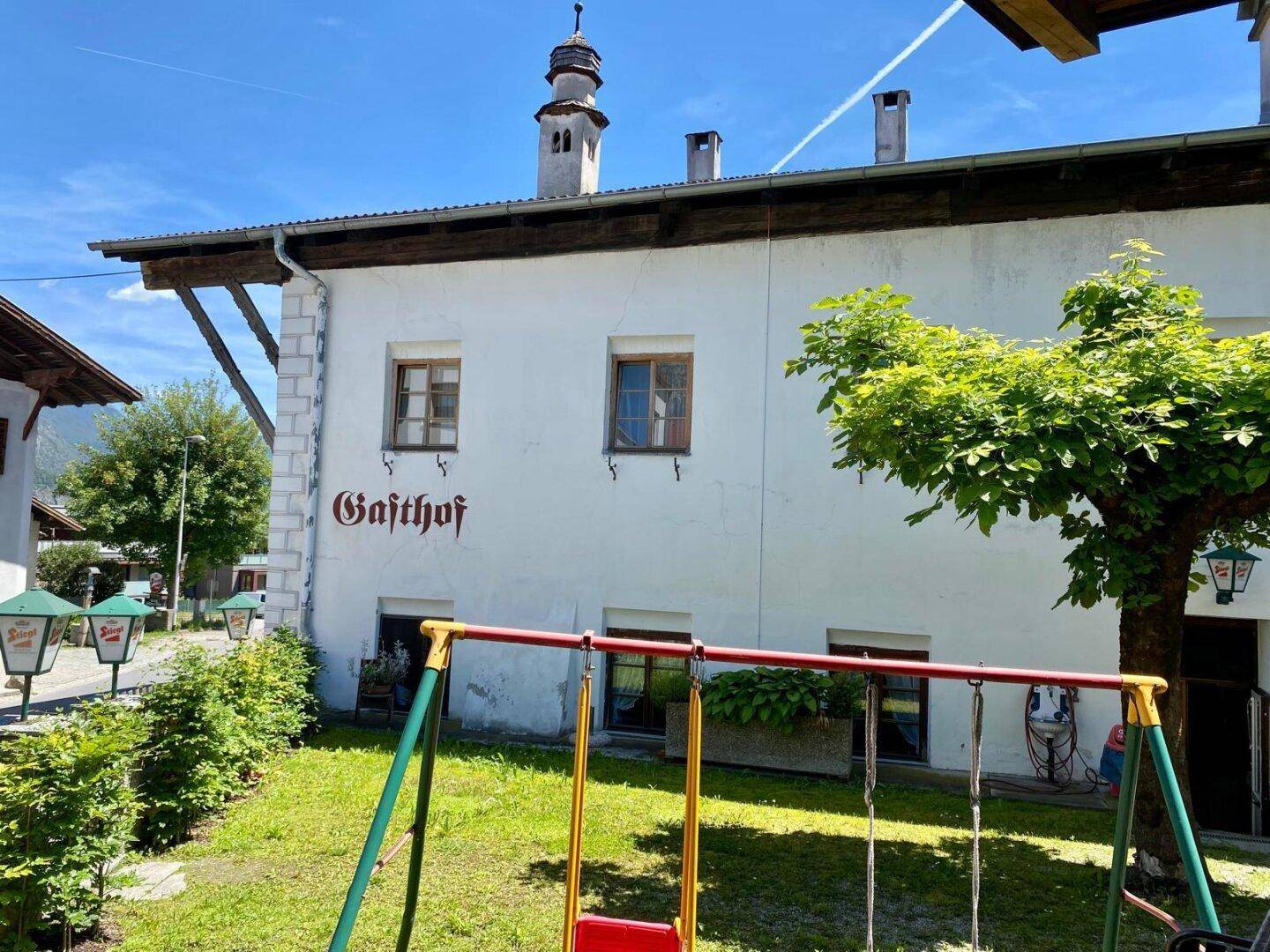 Seitenansicht eines Gasthofs mit Spielplatz im Garten und grüner Umgebung unter blauem Himmel.