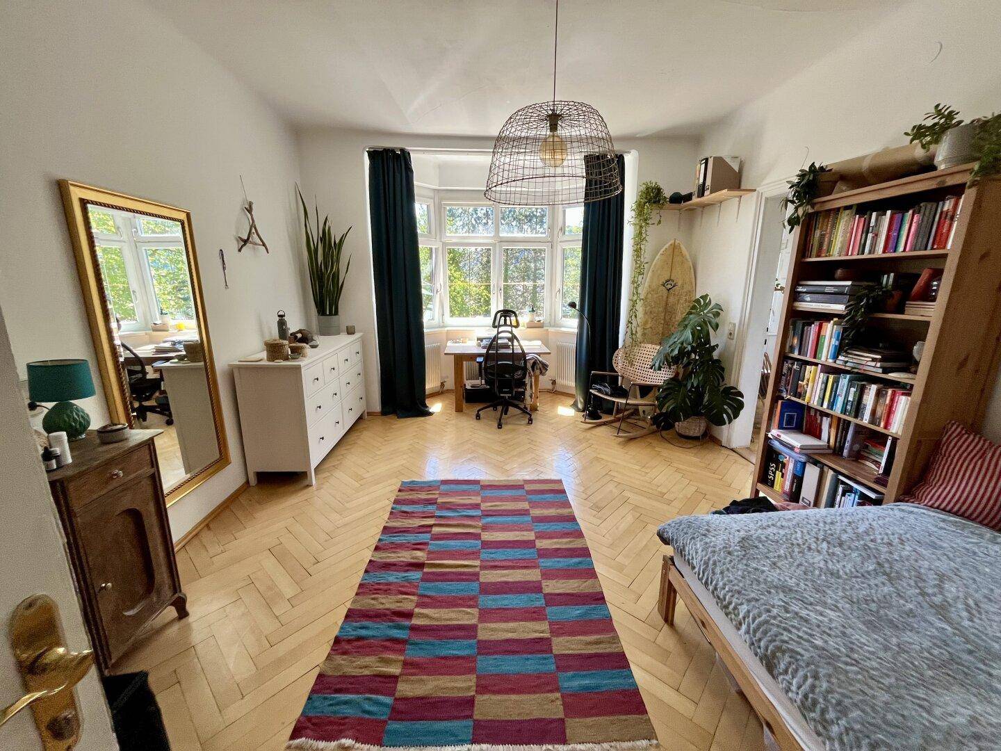 Geräumiges Zimmer mit Parkettboden, großem Erkerfenster und Bücherregal.