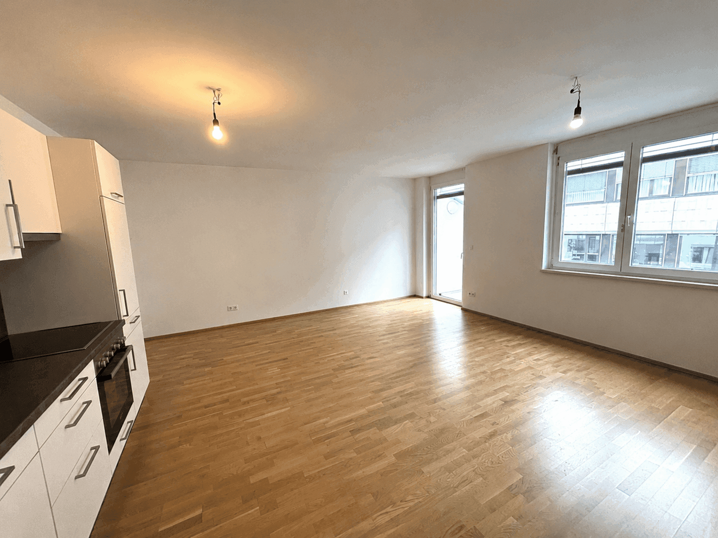Geräumiger, leerer Wohnbereich mit Holzboden, großen Fenstern und Zugang zum Balkon.
