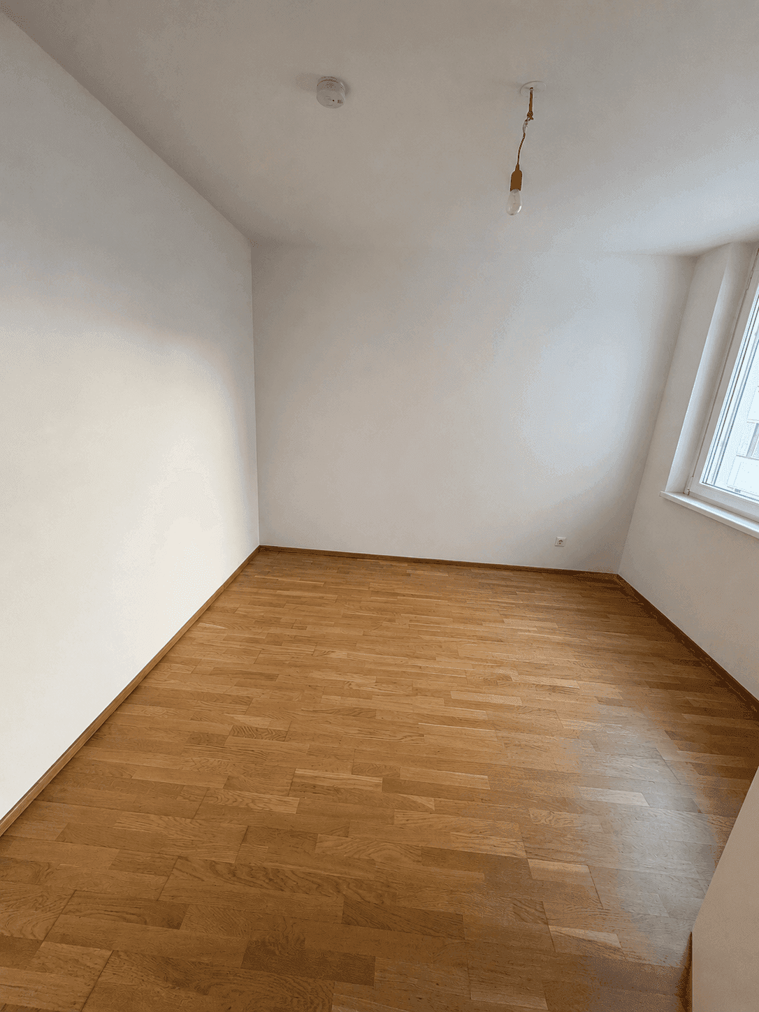 Leeres Zimmer mit Holzboden und einem Fenster, ideal als Schlaf- oder Arbeitszimmer.