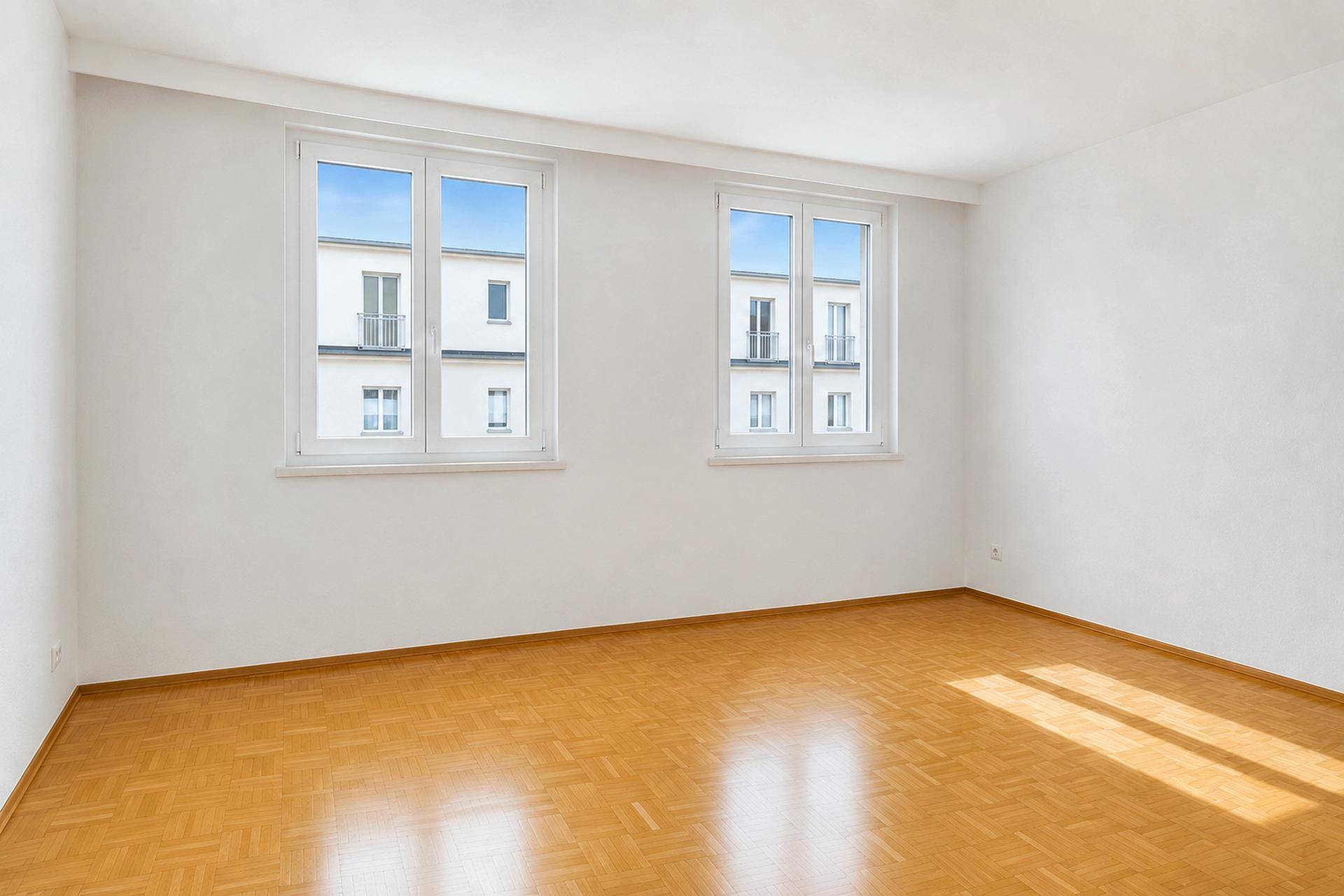 Heller Raum mit Parkettboden und zwei Fenstern, die einen Blick auf die umliegenden Gebäude bieten.