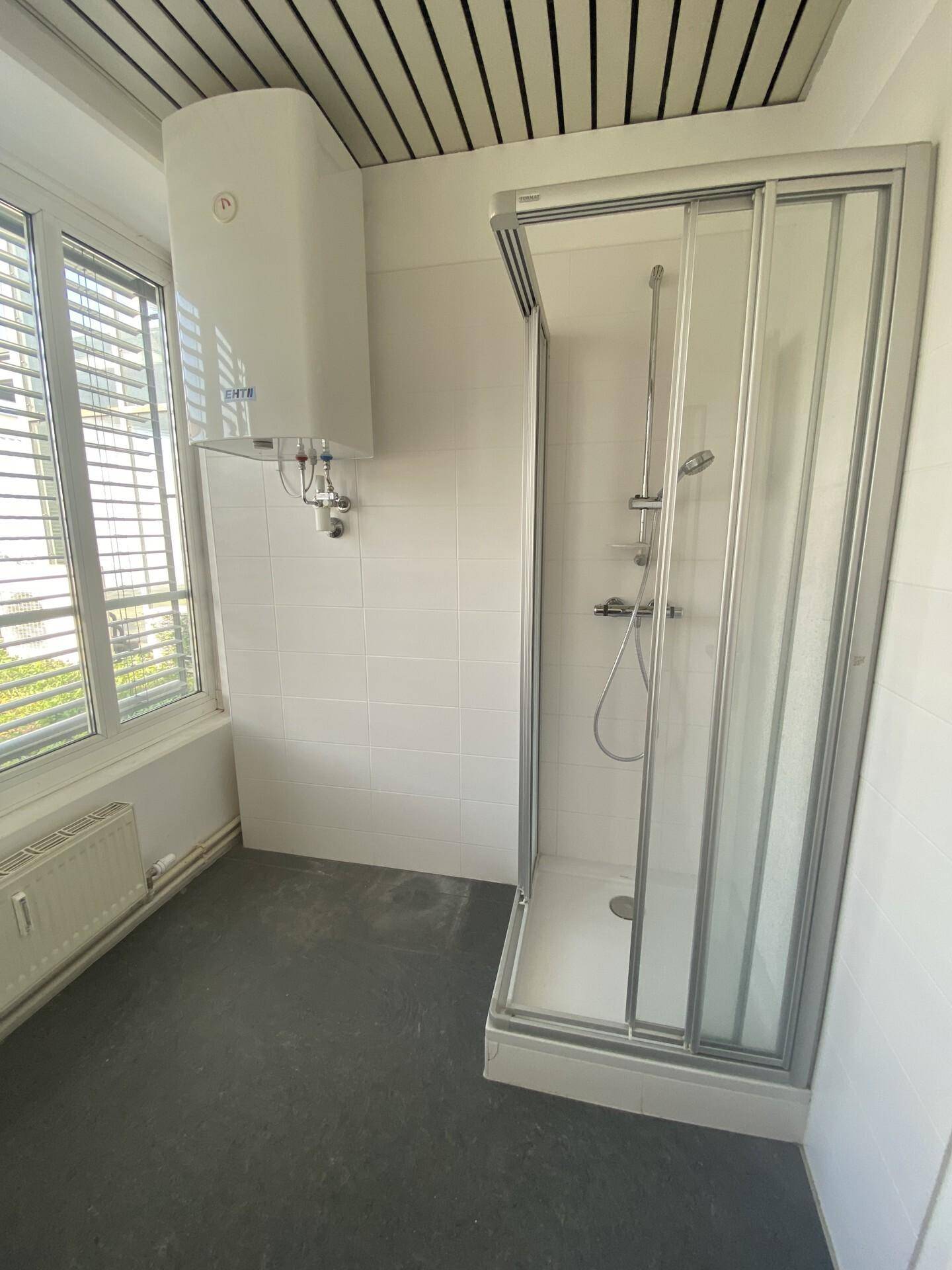 Badezimmer mit Duschkabine, weißen Fliesen, Warmwasserbereiter und einem Fenster mit Jalousien.