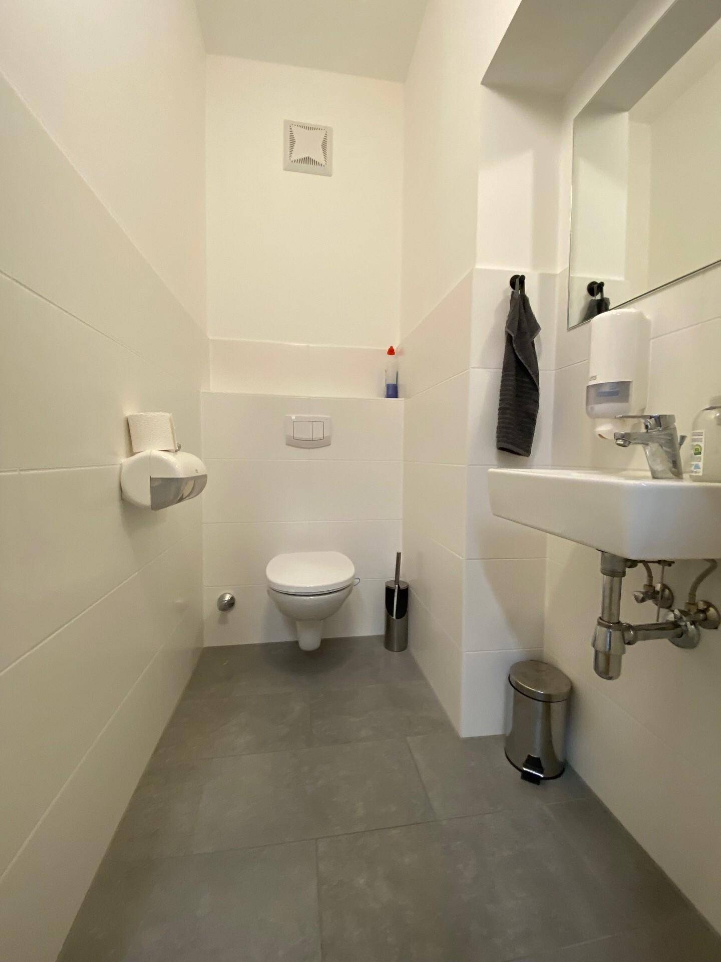Modern geflieste Gästetoilette mit wandhängendem WC, Waschbecken und einem Abluftventilator.