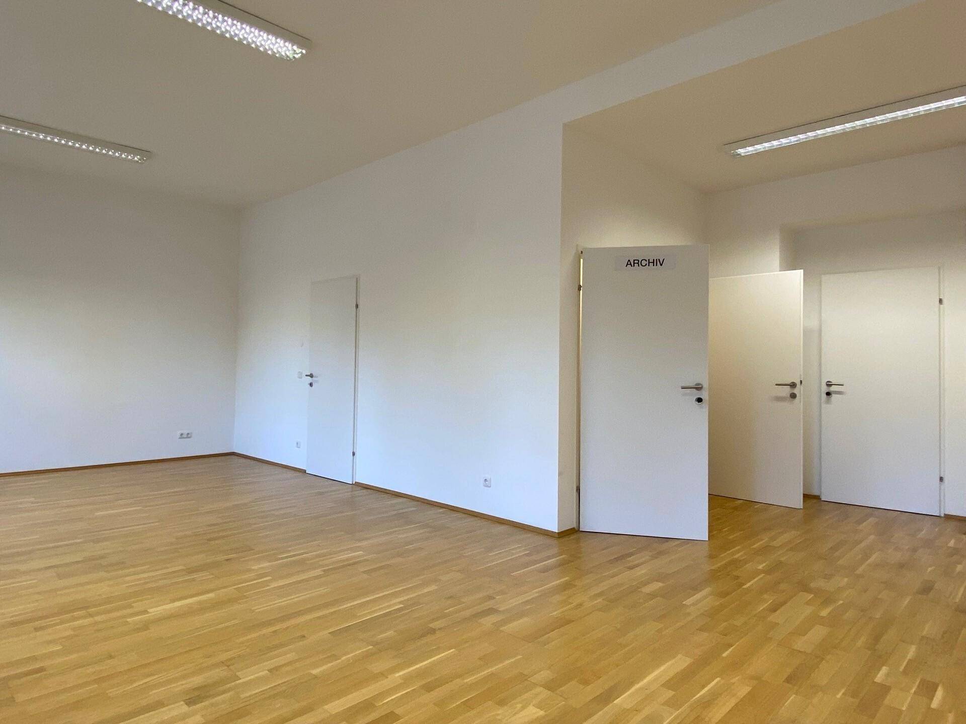 Leerer Büroraum mit hellem Parkettboden und mehreren weißen Türen, eine davon offen.