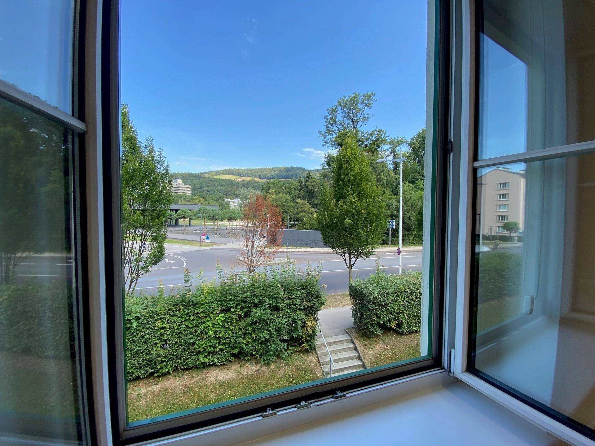 Blick aus dem Fenster auf eine Straße, Bäume und eine hügelige Landschaft im Hintergrund.