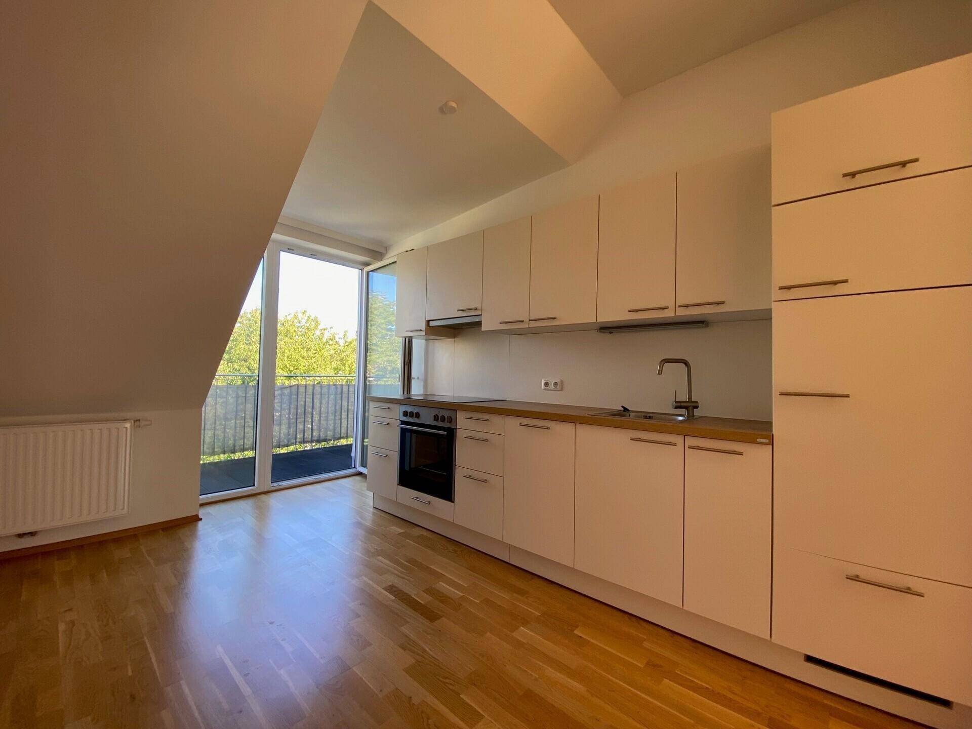 Helle Küche in einer Dachgeschosswohnung mit Einbauschränken, Holzboden und Zugang zum Balkon.
