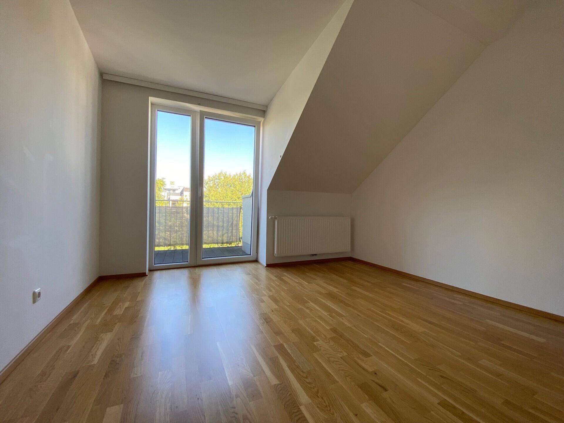 Lichtdurchflutetes Schlafzimmer mit Holzboden, schräger Decke und Balkontür für frische Luft.