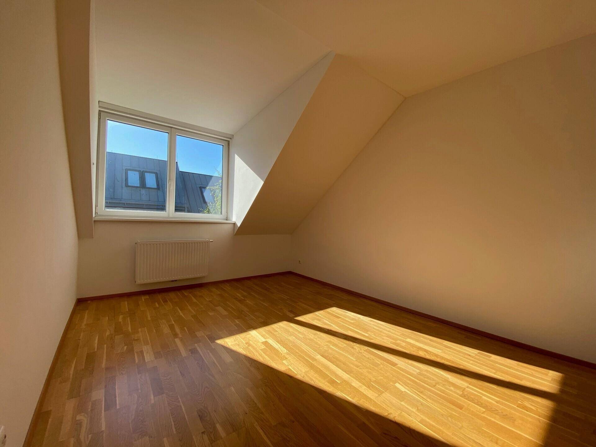 Geräumiges Schlafzimmer mit Holzboden, schräger Decke und großem Fenster für viel Tageslicht.