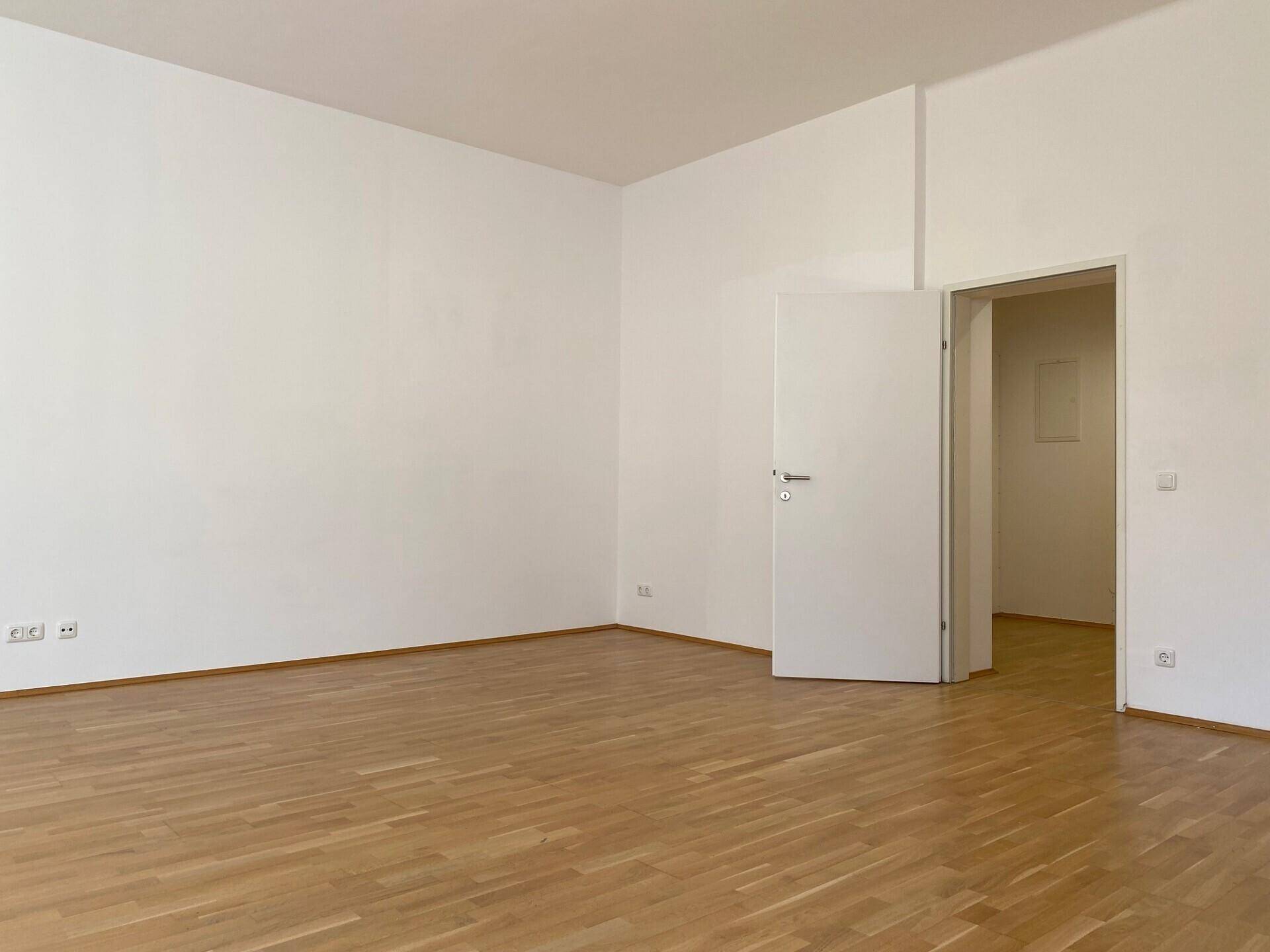 Heller, leerer Raum mit Parkettboden und weißen Wänden, mit Zugang zu einem weiteren Zimmer.