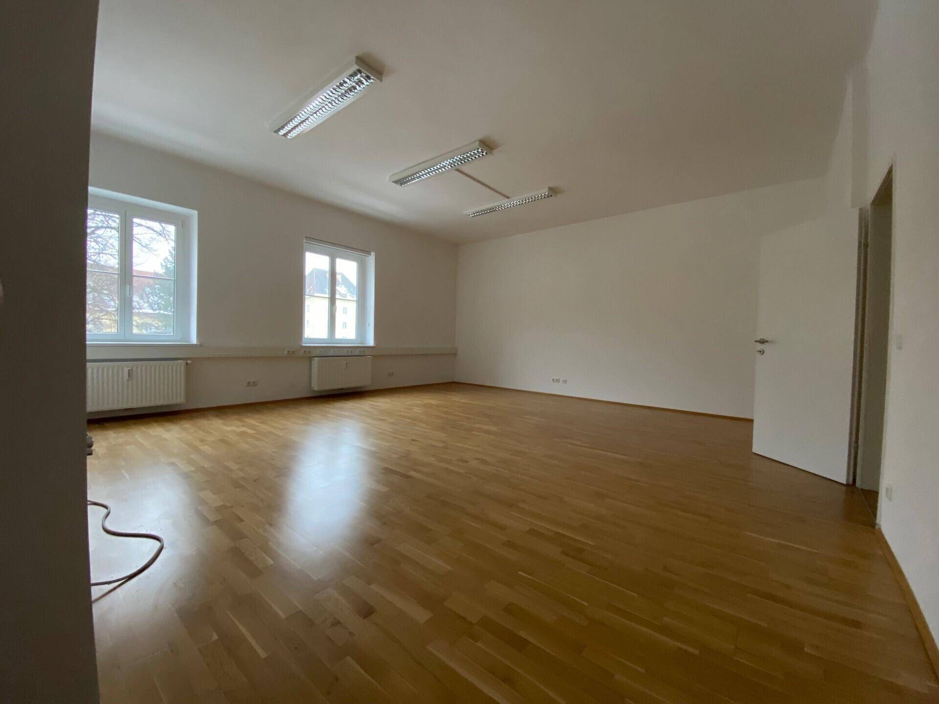 Geräumiger, heller Raum mit Parkettboden und zwei Fenstern, ideal als Wohn- oder Arbeitsbereich.