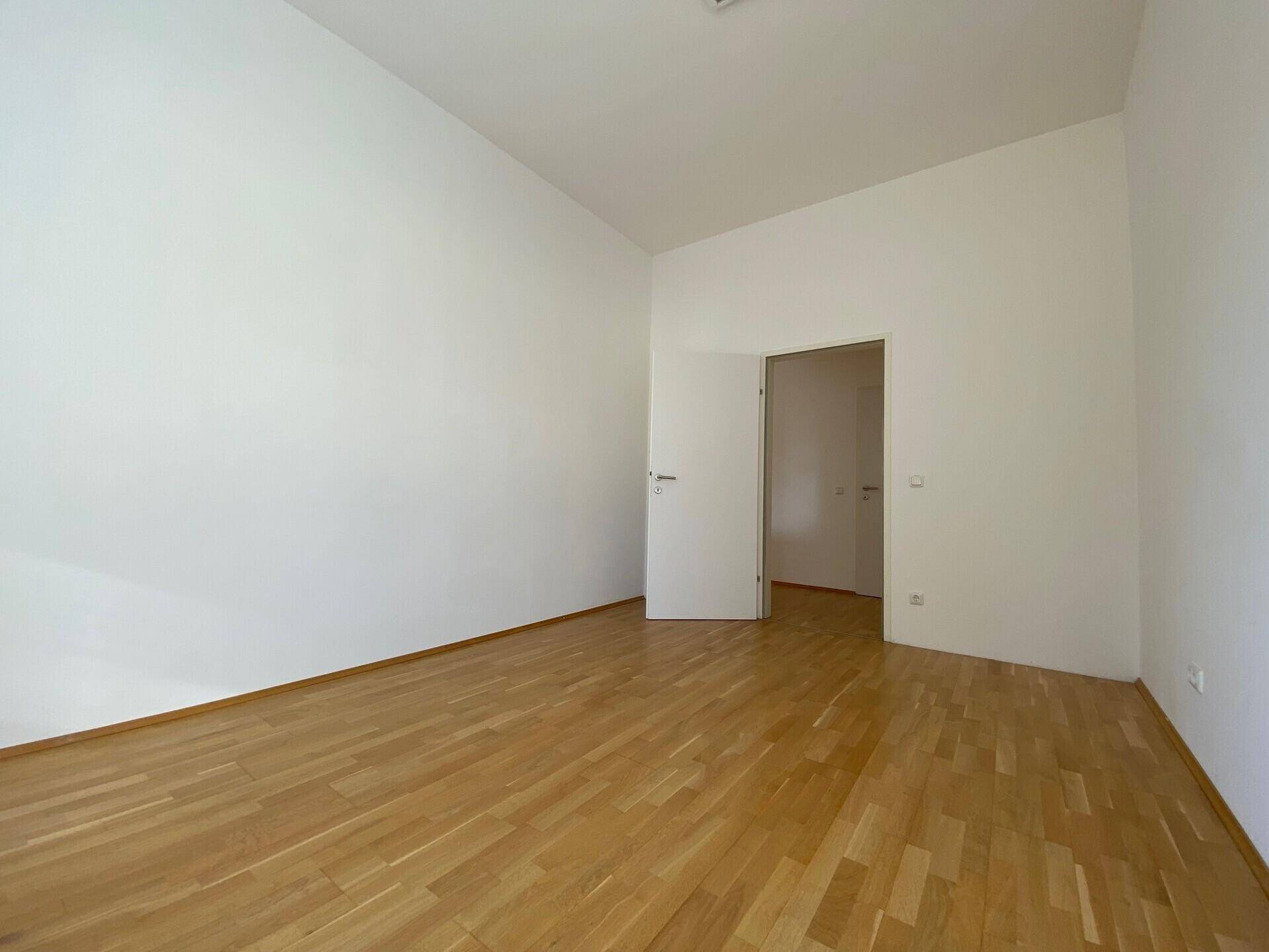 Leeres Zimmer mit Parkettboden und weißen Wänden, bietet Zugang zu einem angrenzenden Raum.
