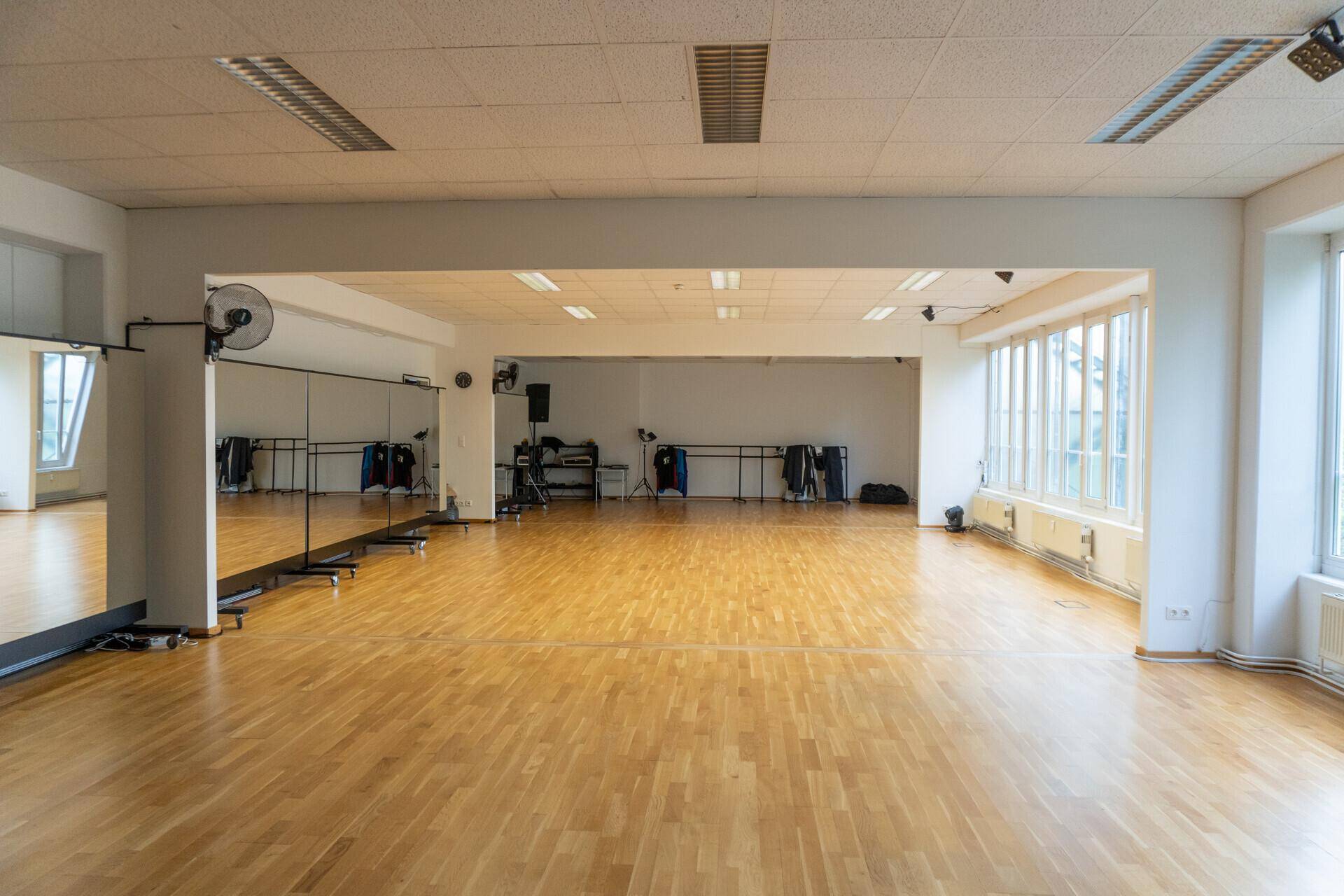 Weitläufiger Studiobereich mit hellem Holzboden, großen Fenstern und Wandspiegeln.