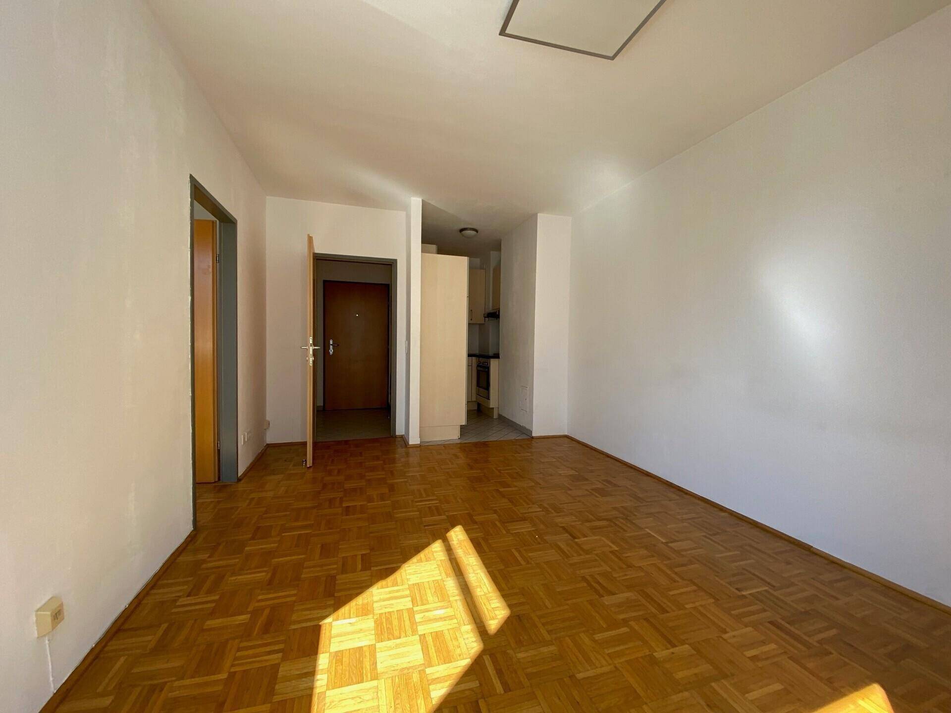 Heller, leerer Wohnraum mit Parkettboden und Zugang zu Flur und Kochnische, durchflutet von Sonnenlicht.