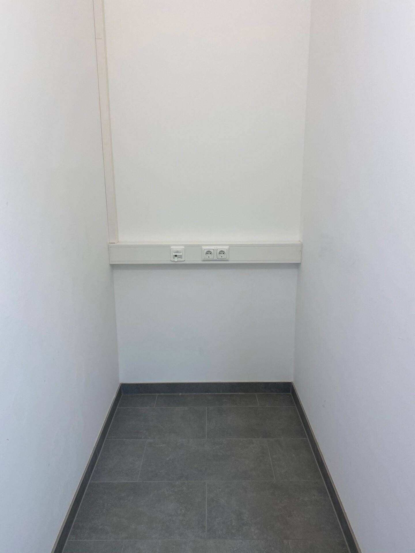 Kleiner, schmaler Raum mit grauem Fliesenboden und weißen Wänden, ausgestattet mit Steckdosen.