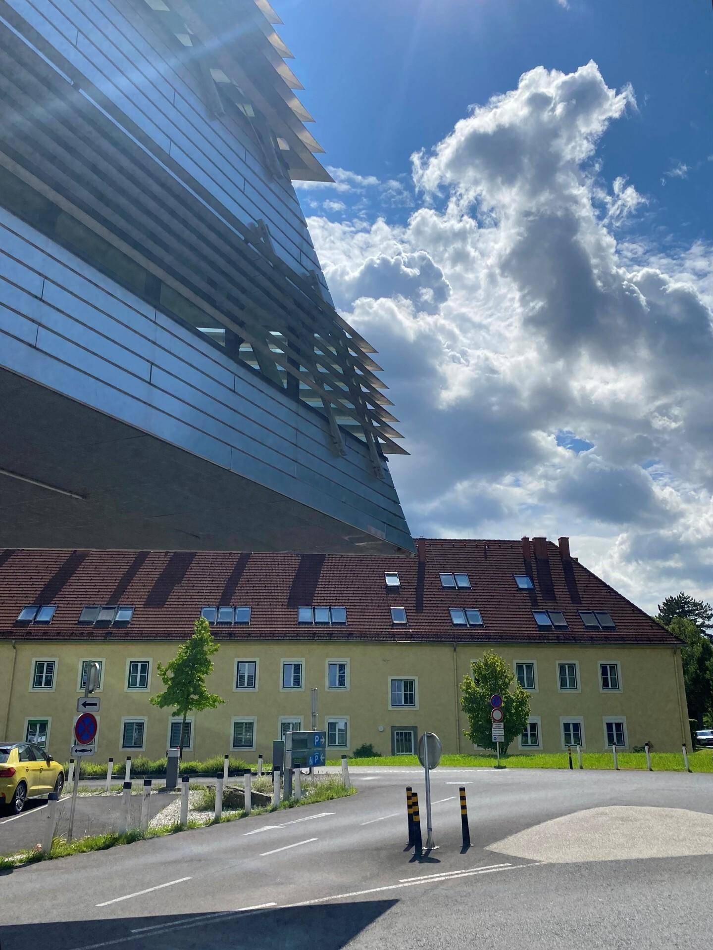Kontrast zwischen moderner Glasfassade und traditionellem Gebäude mit rotem Ziegeldach unter blauem Himmel.