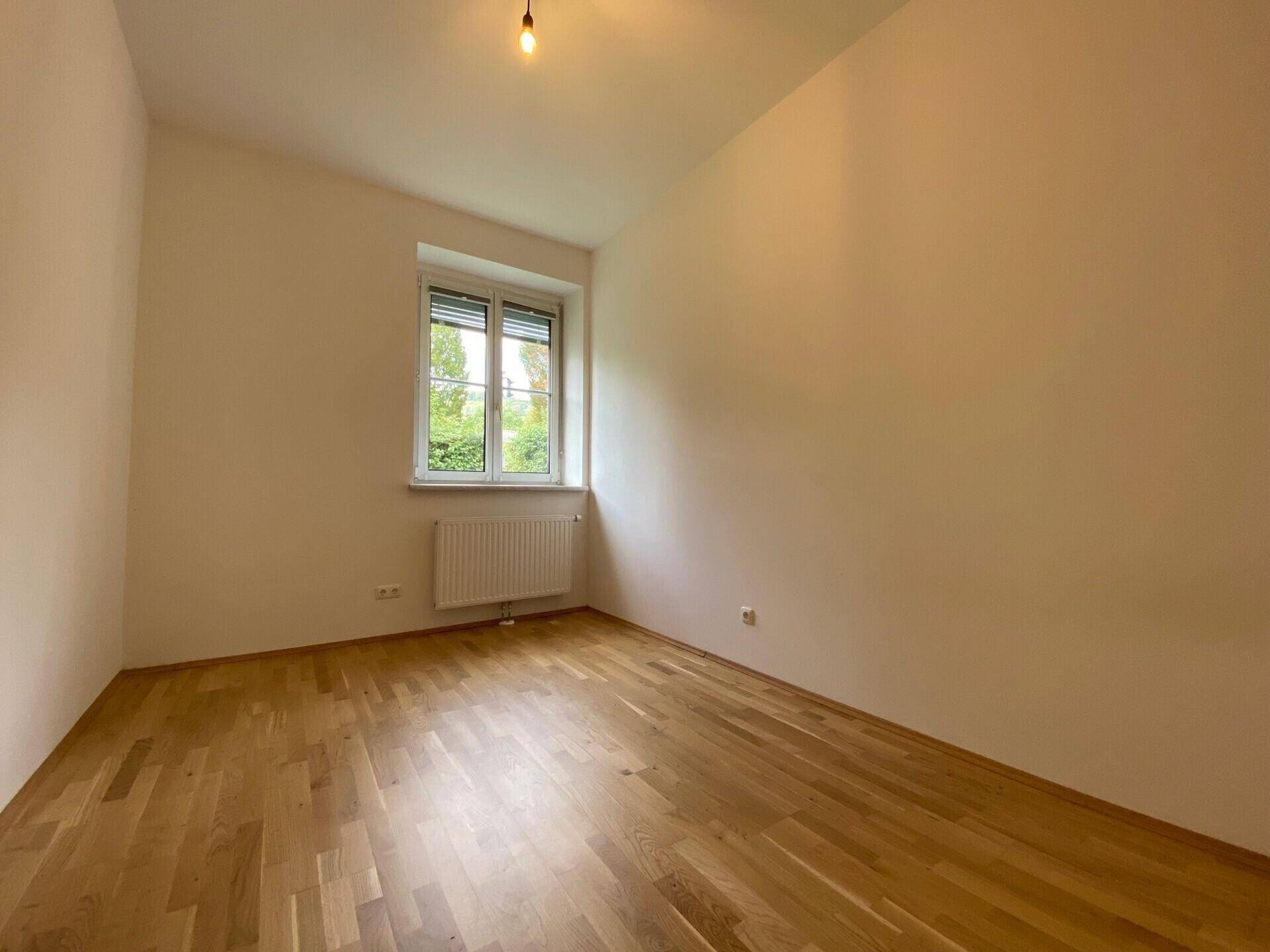 Leeres Zimmer mit Parkettboden, weiß gestrichenen Wänden und einem Fenster mit Blick ins Grüne.