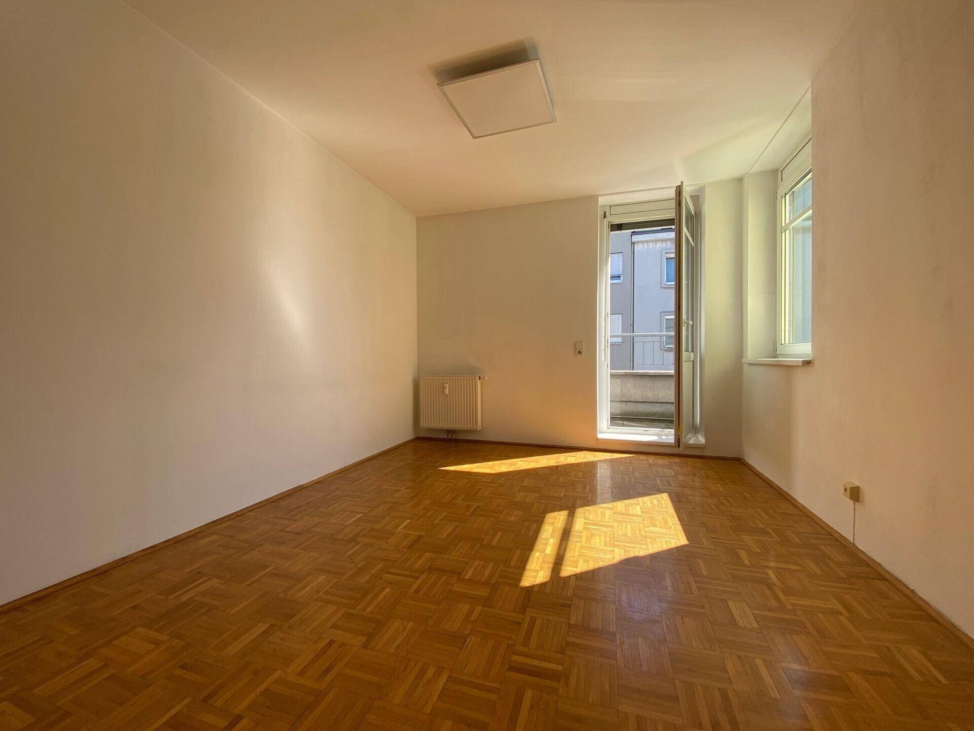 Lichtdurchfluteter Raum mit Parkettboden, Heizkörper und Zugang zu einem kleinen Balkon, ideal für Wohnzwecke.