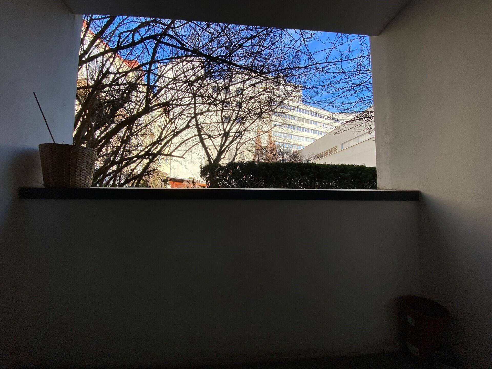 Blick von einer Loggia auf kahle Bäume und ein entferntes, modernes Gebäude unter strahlend blauem Himmel.