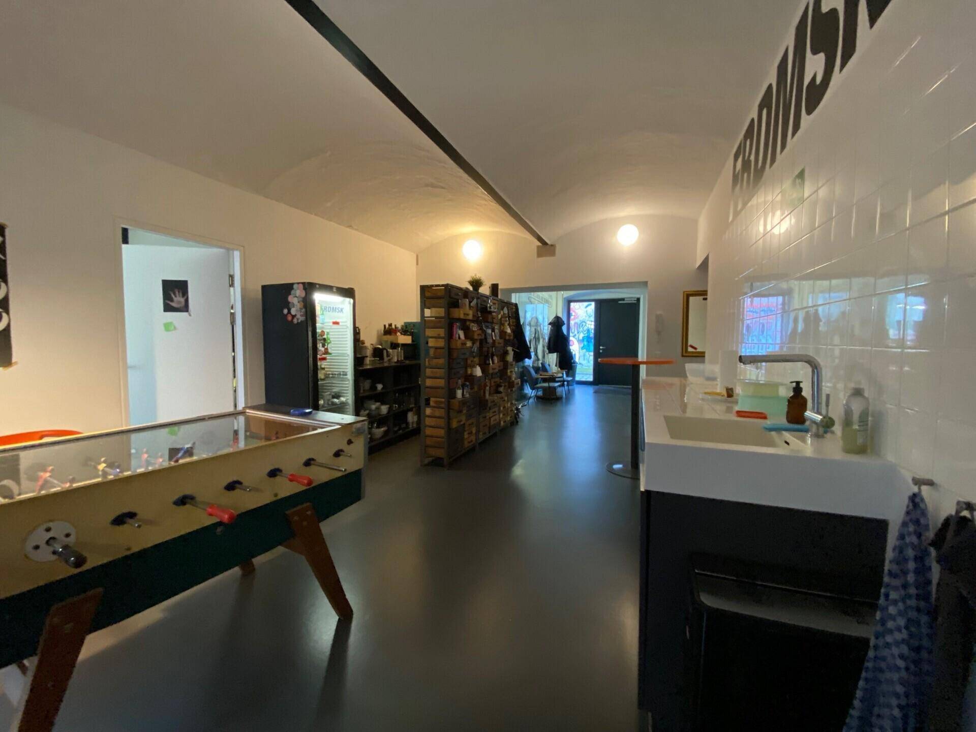 Offener Bürobereich mit Kickertisch, kleiner Küchenzeile und direktem Zugang zum Außenbereich unter einer gewölbten Decke.
