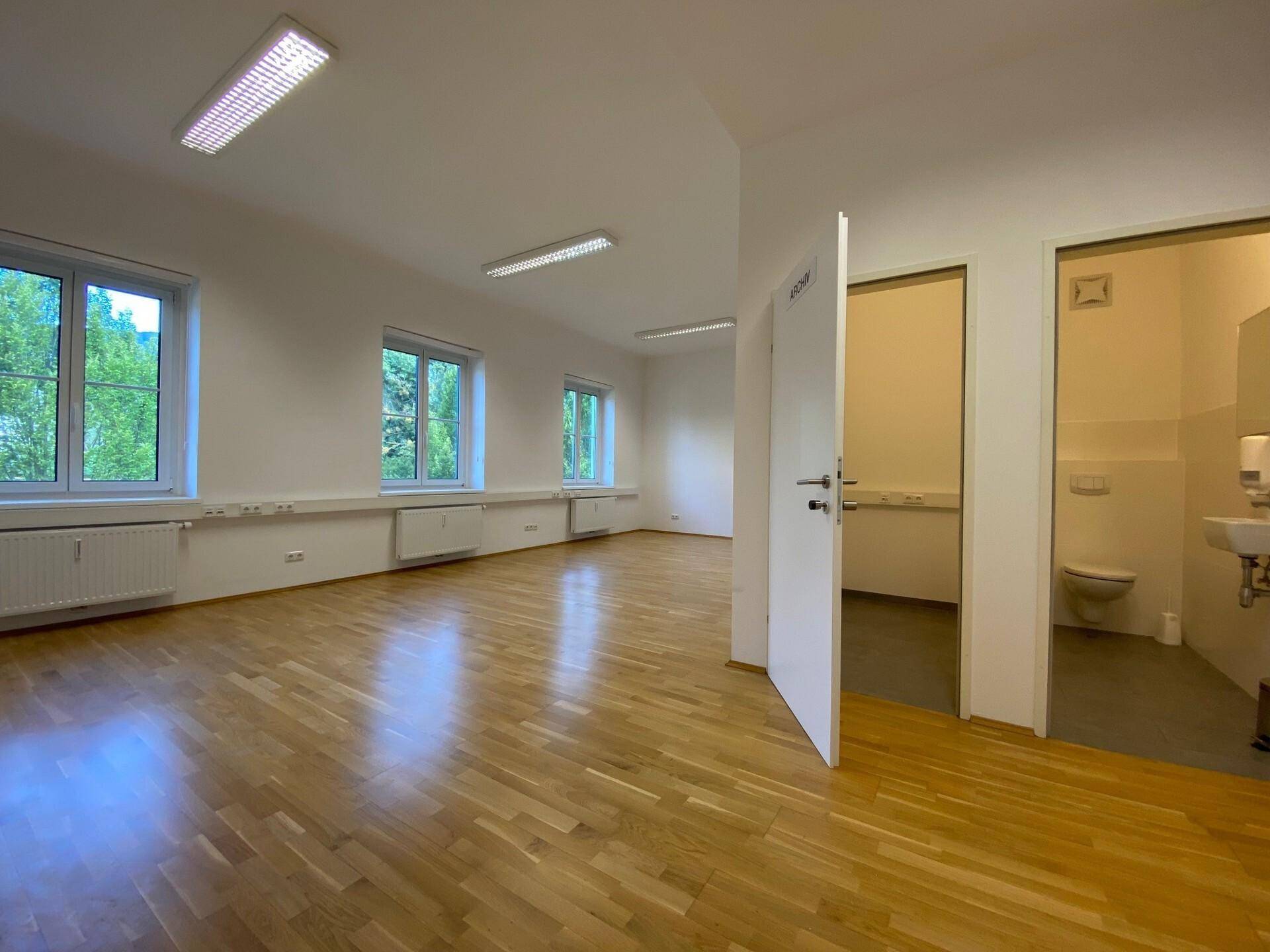 Heller Büroraum mit Parkettboden, geöffneten Türen zu Archiv und WC, sowie großen Fenstern.