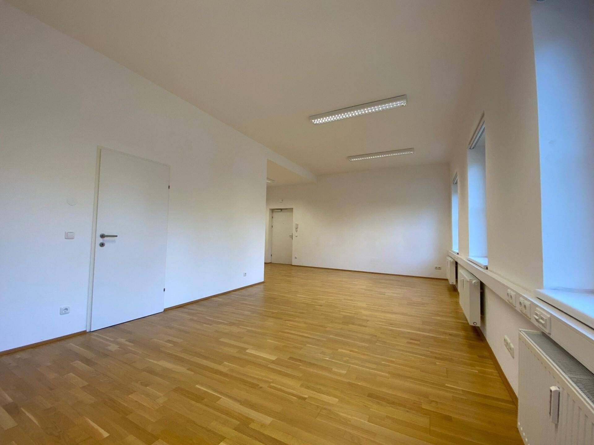 Geräumiger Büroraum mit Parkettboden, weißen Wänden, zwei Fenstern und einer weißen Tür.