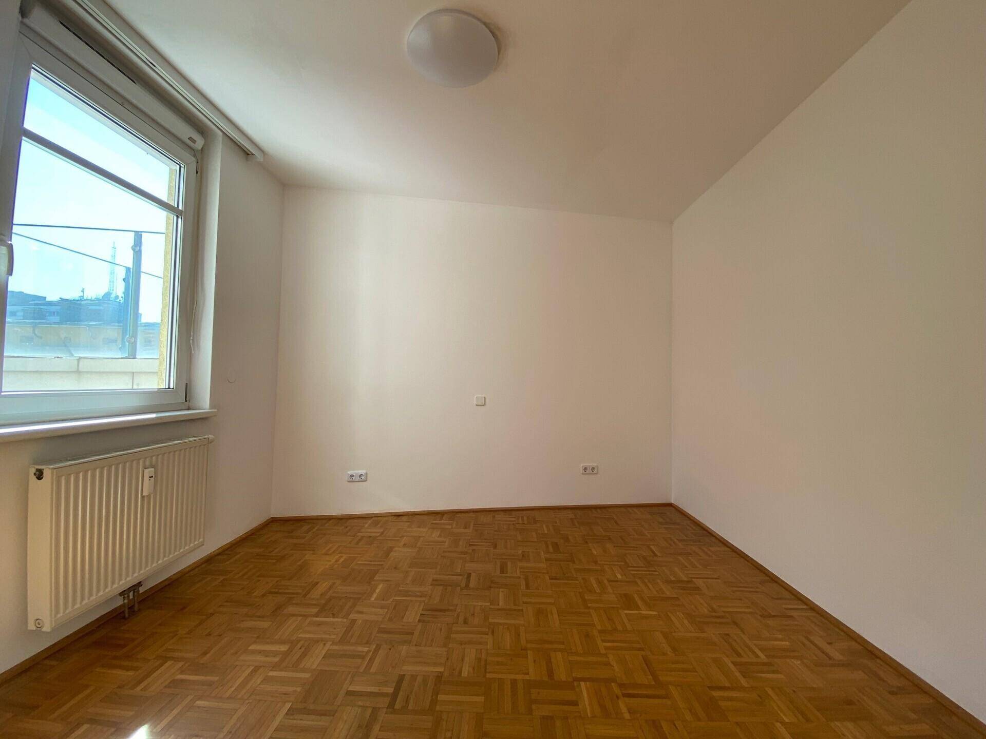 Helles Schlafzimmer mit Parkettboden, weiß gestrichenen Wänden und großem Fenster für viel Tageslicht.