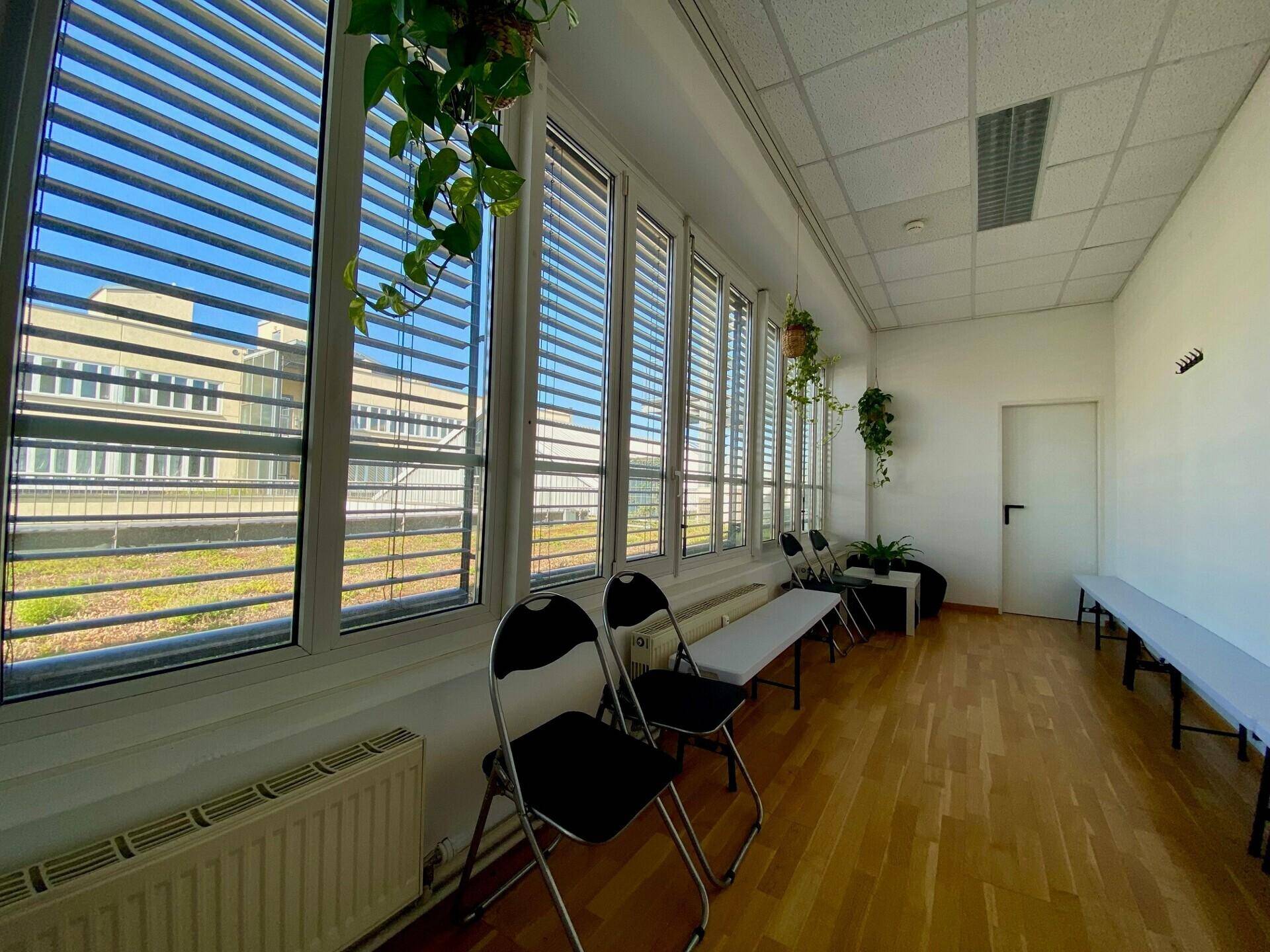 Heller Wartebereich mit Holzboden, großen Fenstern mit Jalousien und einfachen Sitzgelegenheiten.