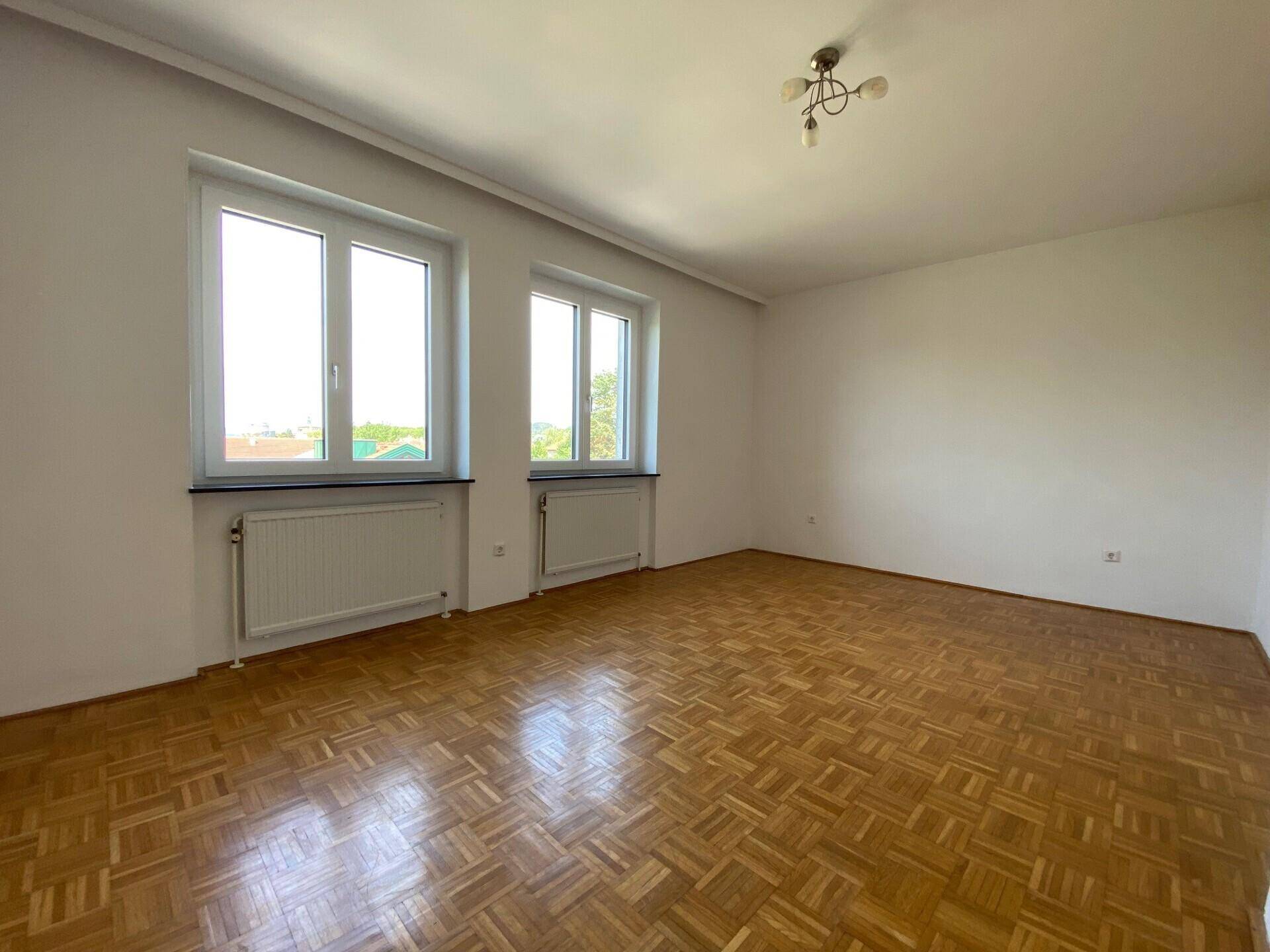 Heller Raum mit klassischem Parkettboden und zwei großen Fenstern für viel Tageslicht.