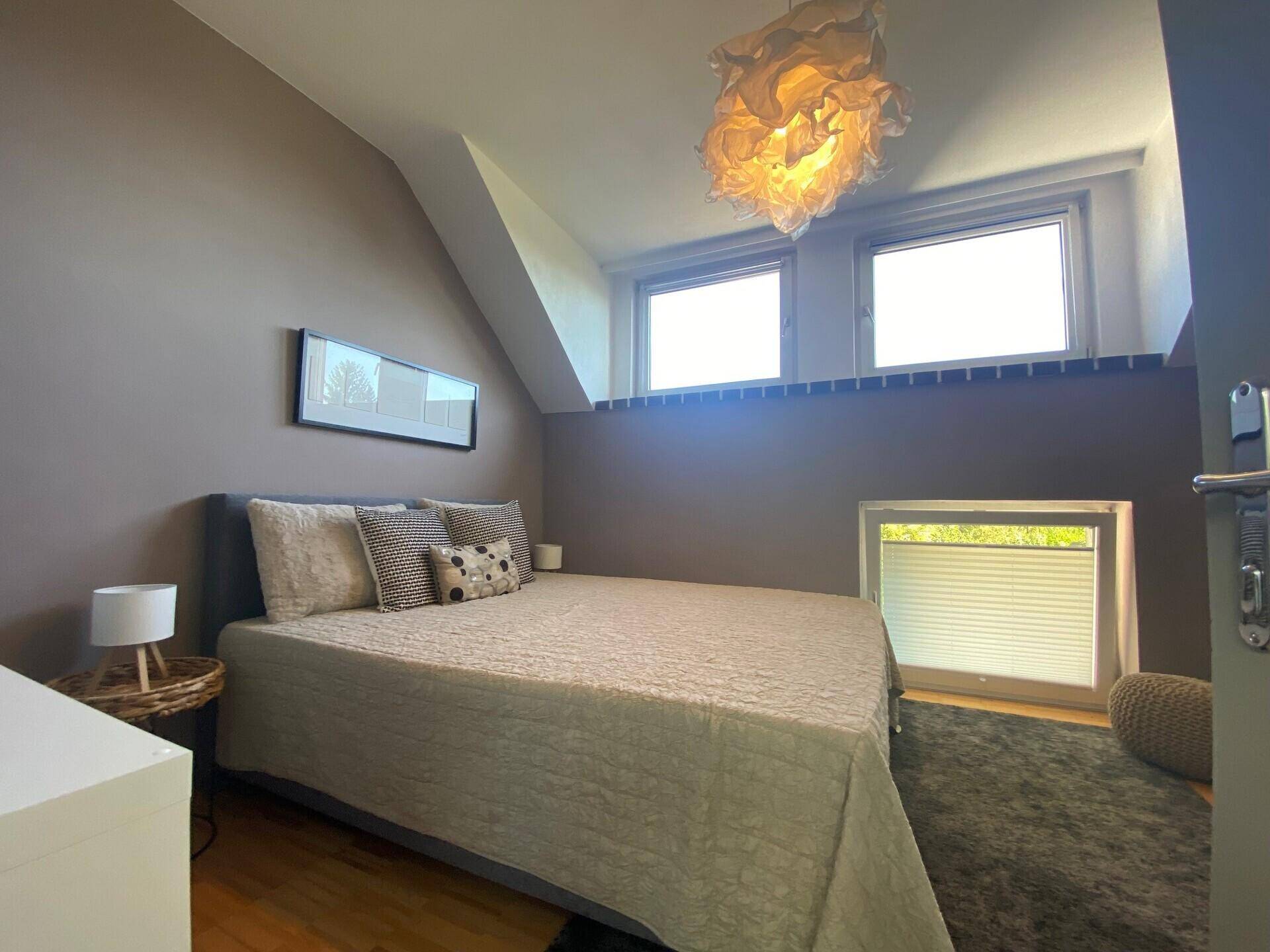 Helles Schlafzimmer mit Doppelbett, Holzboden und Fenstern, die viel Tageslicht hereinlassen.