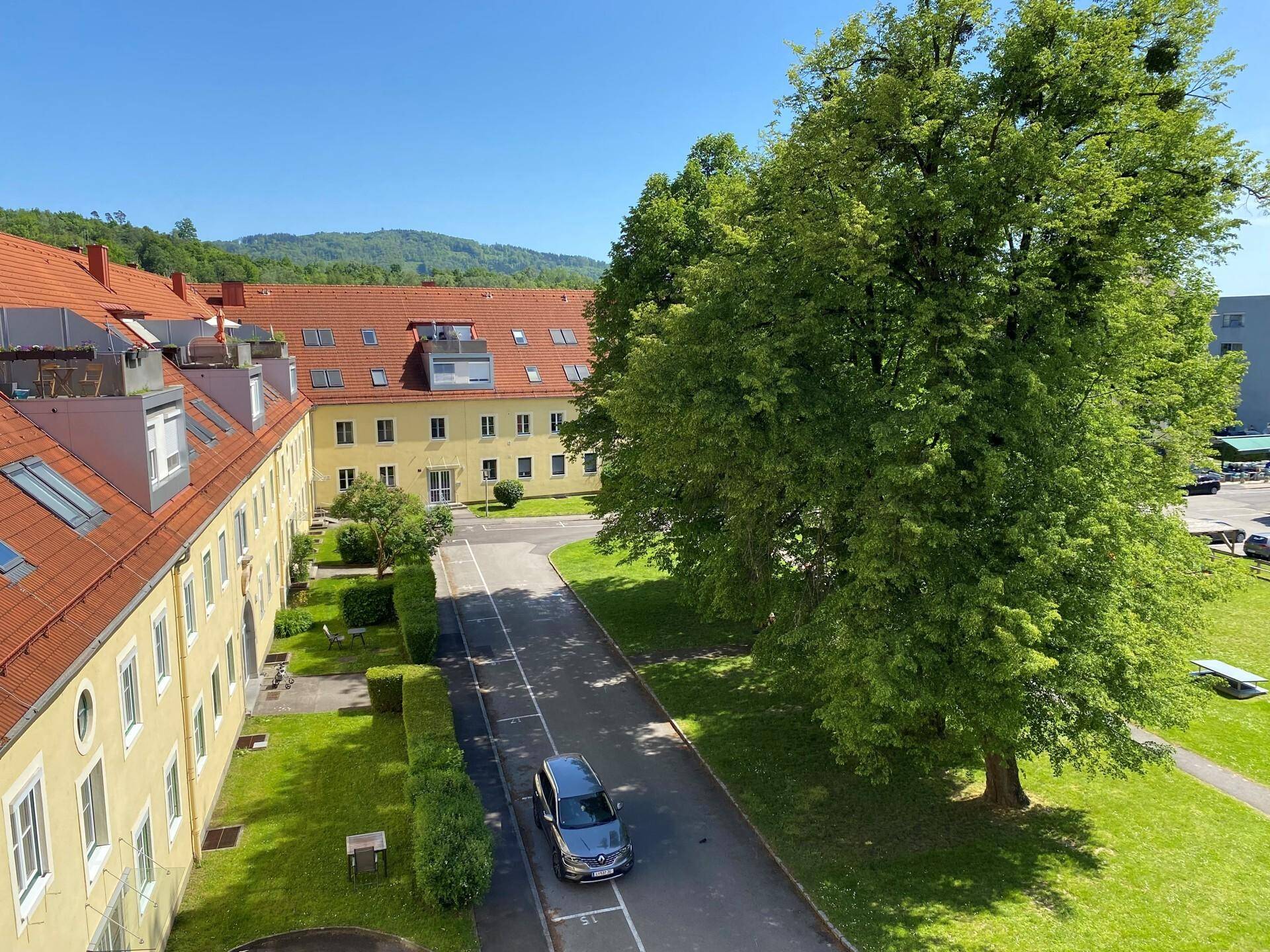 Luftaufnahme eines Gebäudekomplexes mit roten Dächern, viel Grünfläche und Bäumen unter blauem Himmel.